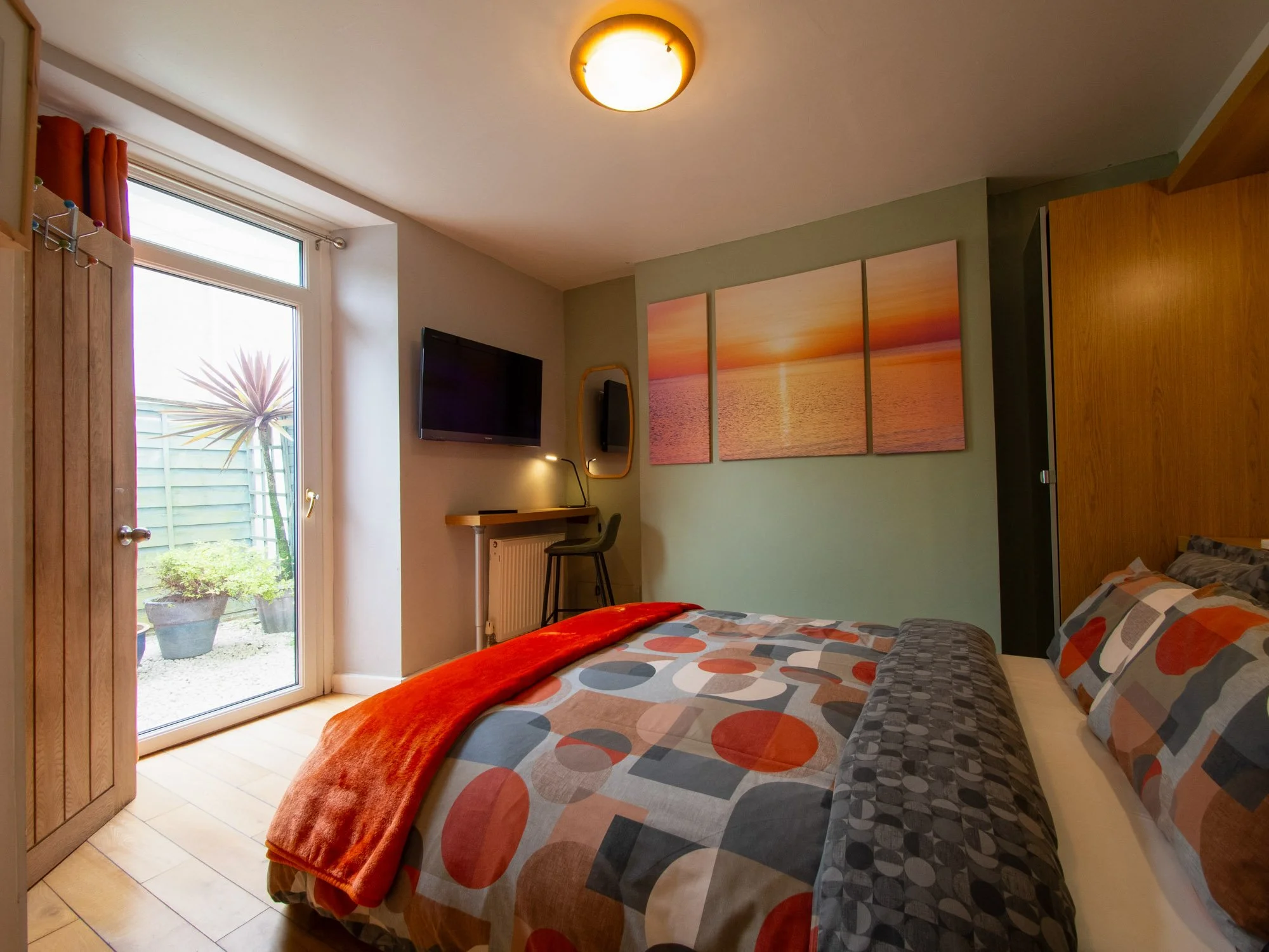 A bedroom with a double bed covered in a patterned duvet and pillows, a red blanket at the foot, a wall-mounted flat-screen TV, and a large painting of a sunset over water. There's a glass door opening to a garden with a potted plant outside.