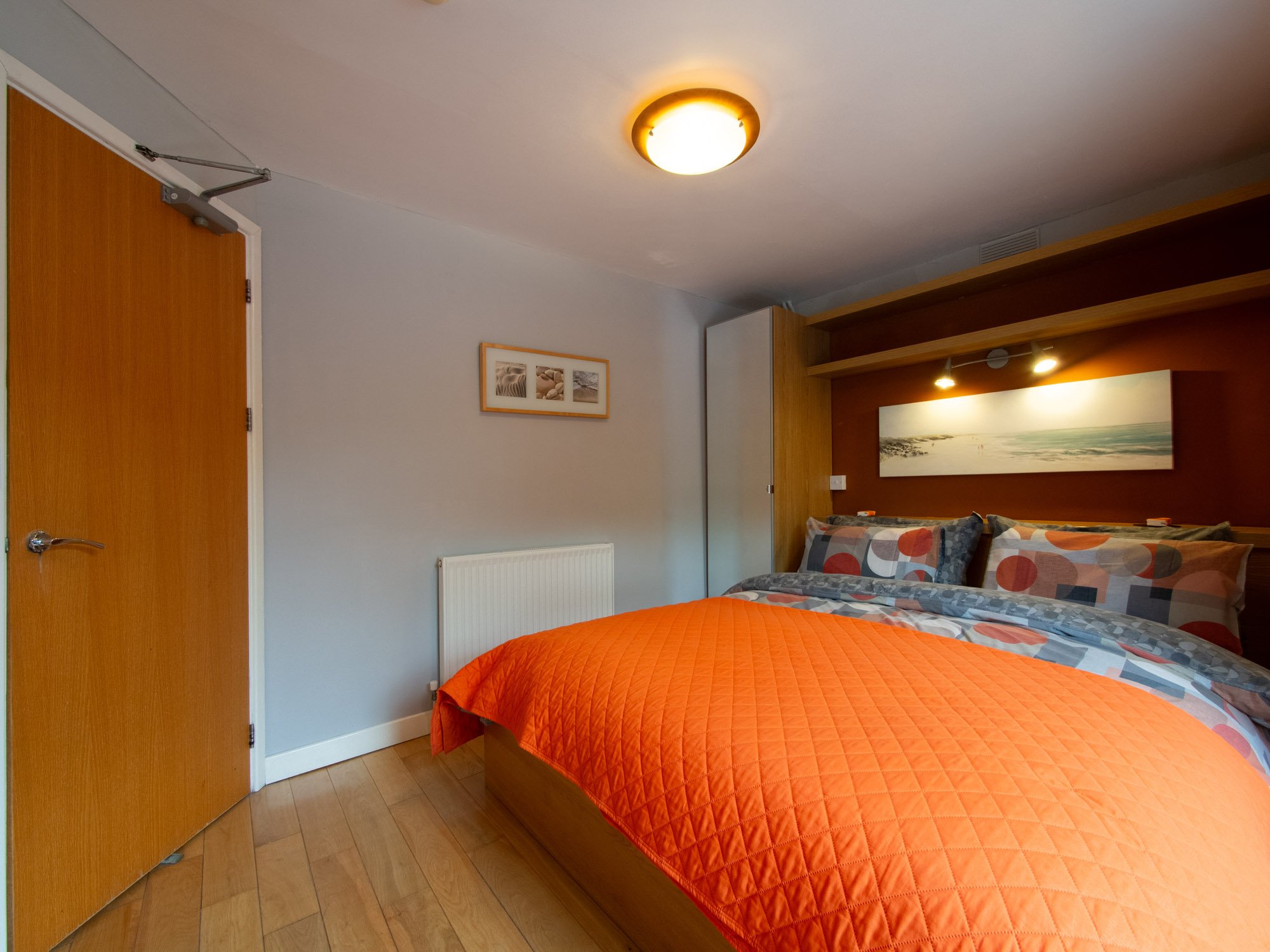 A bedroom with a wooden double bed covered with an orange quilt and patterned pillows. The wall above the bed has a wide beach scene painting, and smaller framed beach-themed prints are on the adjacent wall. The room has a wooden door on the left, a 
