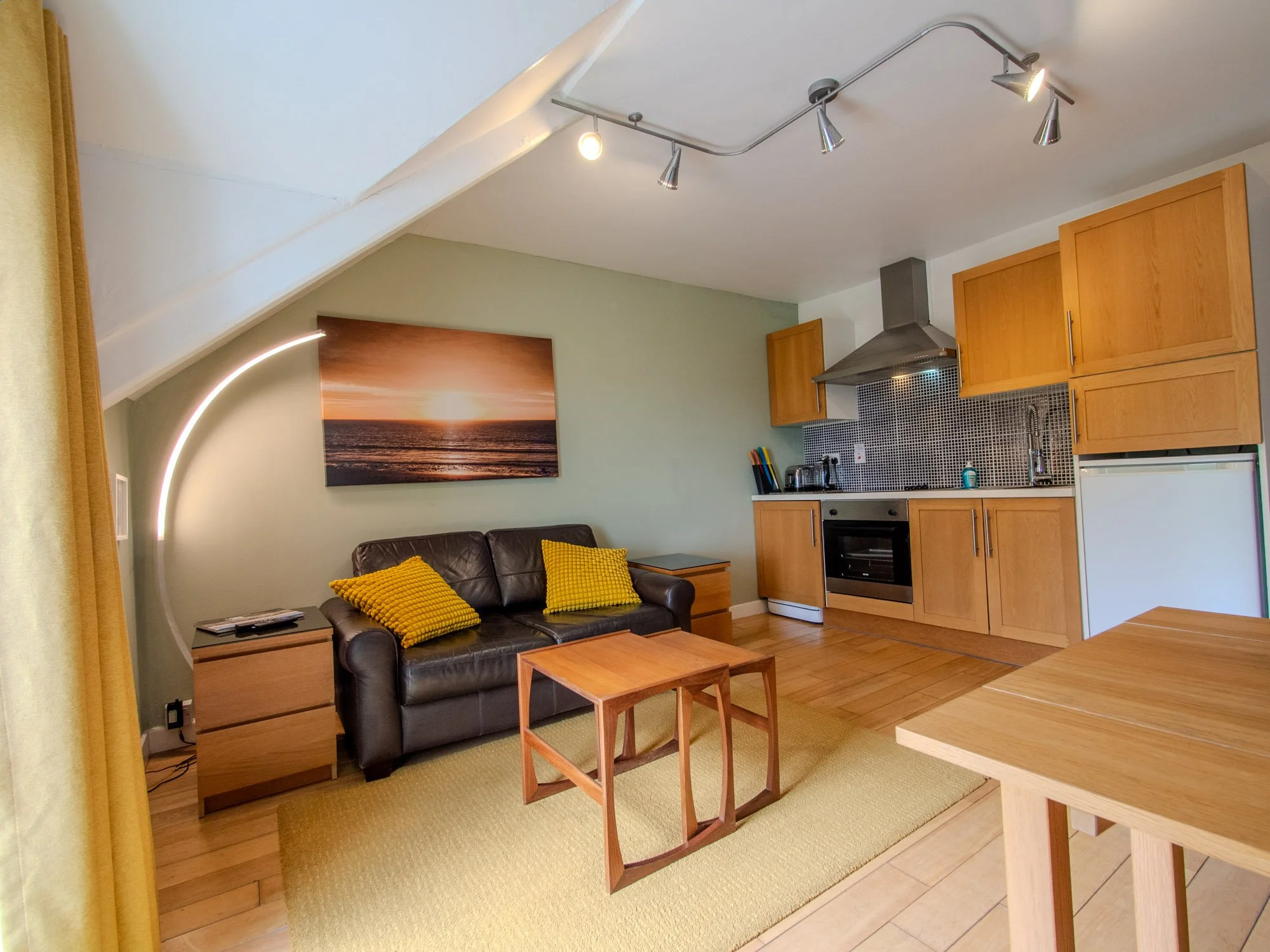 A cozy living area with a black leather sofa with yellow pillows, a wooden side table, a sunset ocean art piece on the green wall, and a kitchenette with wooden cabinets, black tile backsplash, and stainless steel appliances under ceiling track light