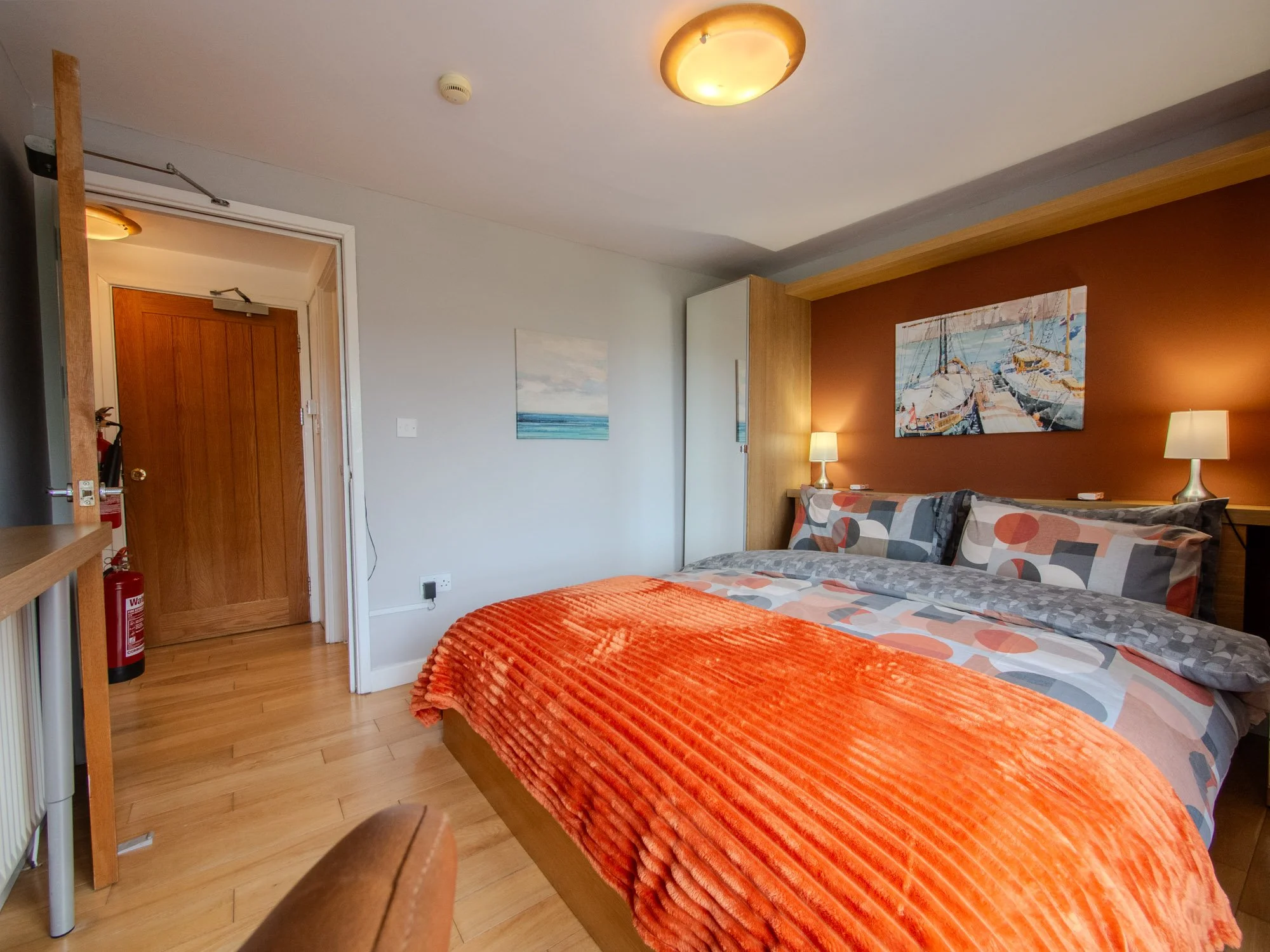 A cozy bedroom with a bed covered in a red textured blanket and pillows featuring geometric patterns. There are two bedside lamps on either side of the bed. The wall behind the bed is painted reddish-brown and decorated with a nautical-themed paintin
