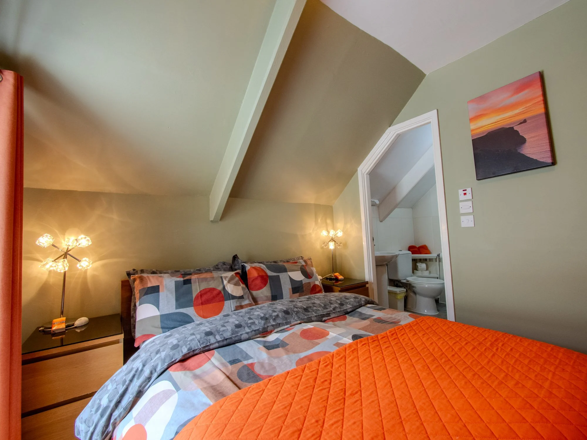 A bedroom with a bed that has a colorful geometric pattern bedspread and pillows. Next to the bed is a black nightstand with a lamp, alarm clock, and remote. There is a small bathroom with a toilet, sink, and mirror visible through an open door. The 