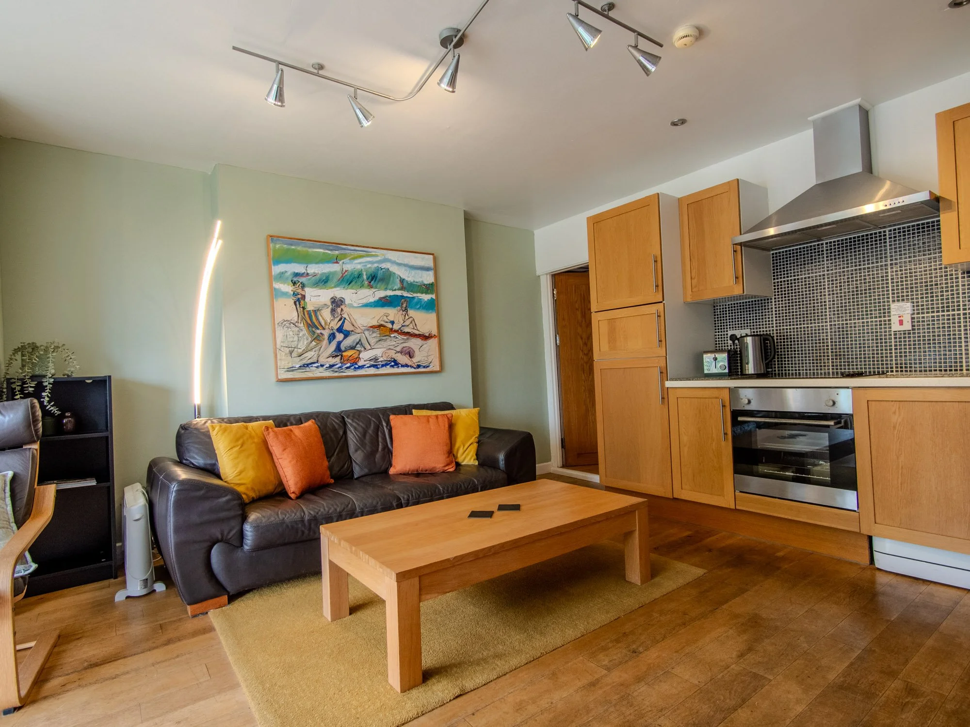 Living room with leather sofa, colorful pillows, wooden coffee table, artwork of a beach scene on the wall, and kitchen area with wooden cabinets, black backsplash, and stainless steel appliances.