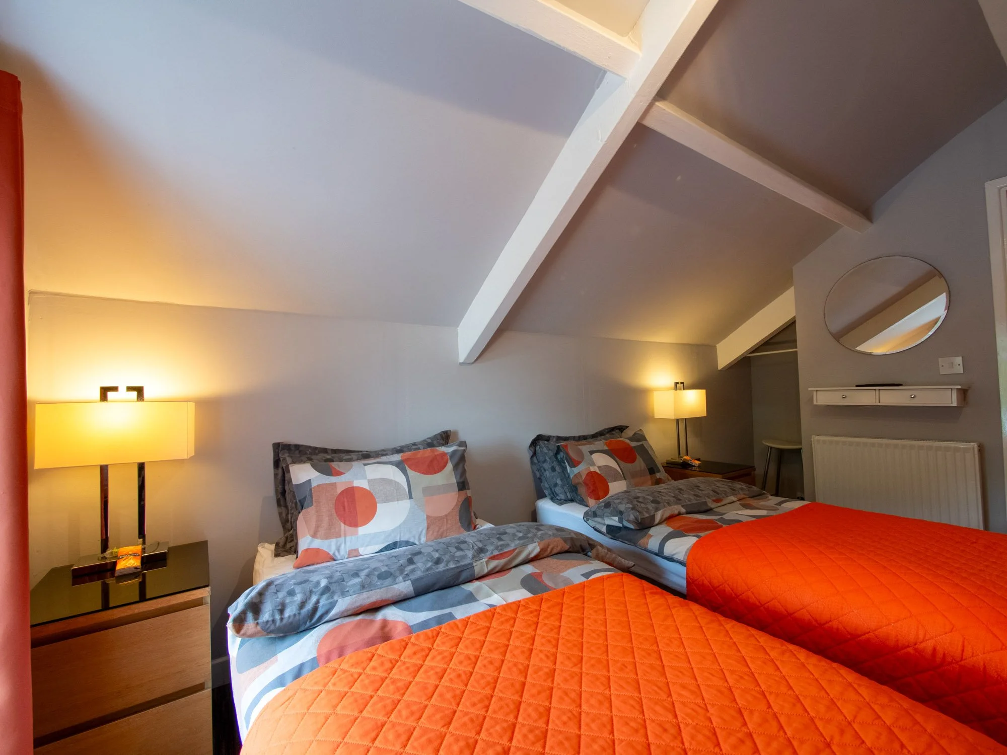 A bedroom with two beds, decorated in gray and orange bedding. There are two pillows on each bed with a geometric pattern. A wooden nightstand with a black tabletop holds a lamp with a rectangular shade and a small object. The wall behind the beds is