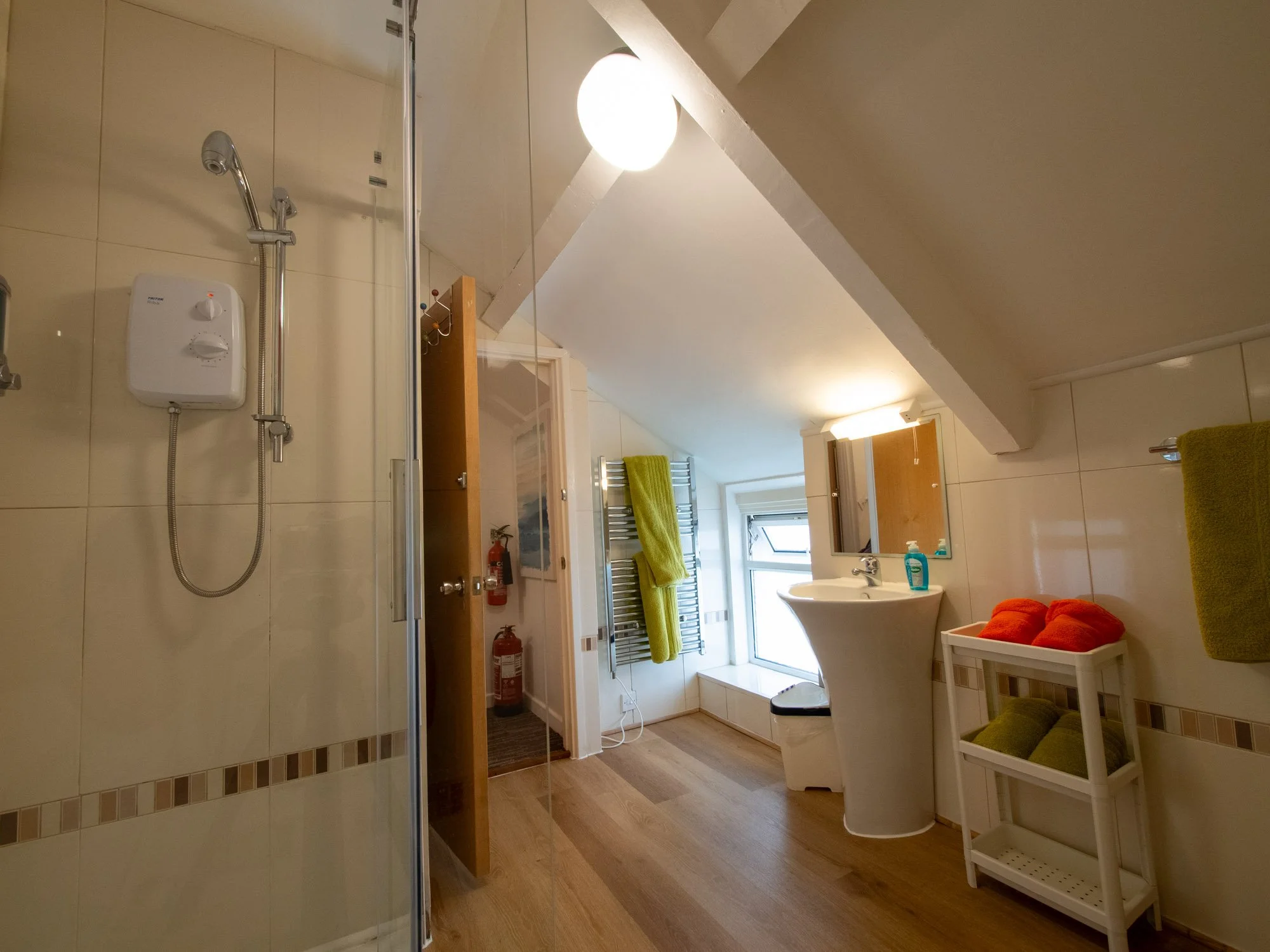 A bathroom with a shower on the left, a sink with a mirror on the right, a towel rack with green towels, wooden cabinets above the sink, a white washbasin, a window with a open sash, and a wooden floor.