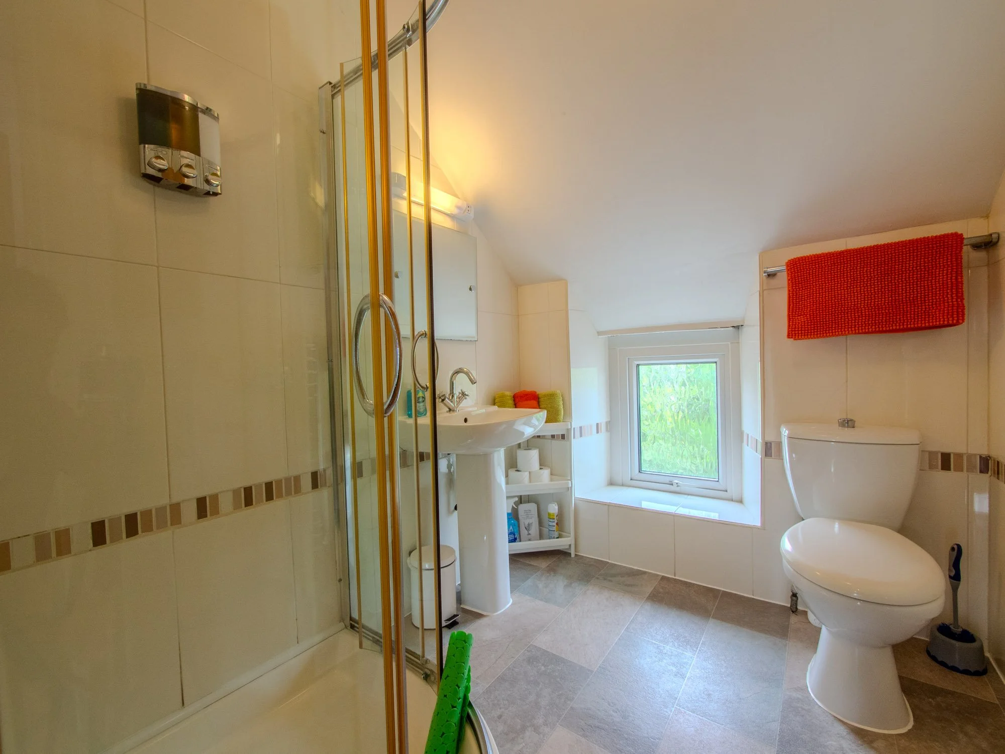 A bathroom with a shower stall, a sink, a toilet, a window, and some colorful towels.