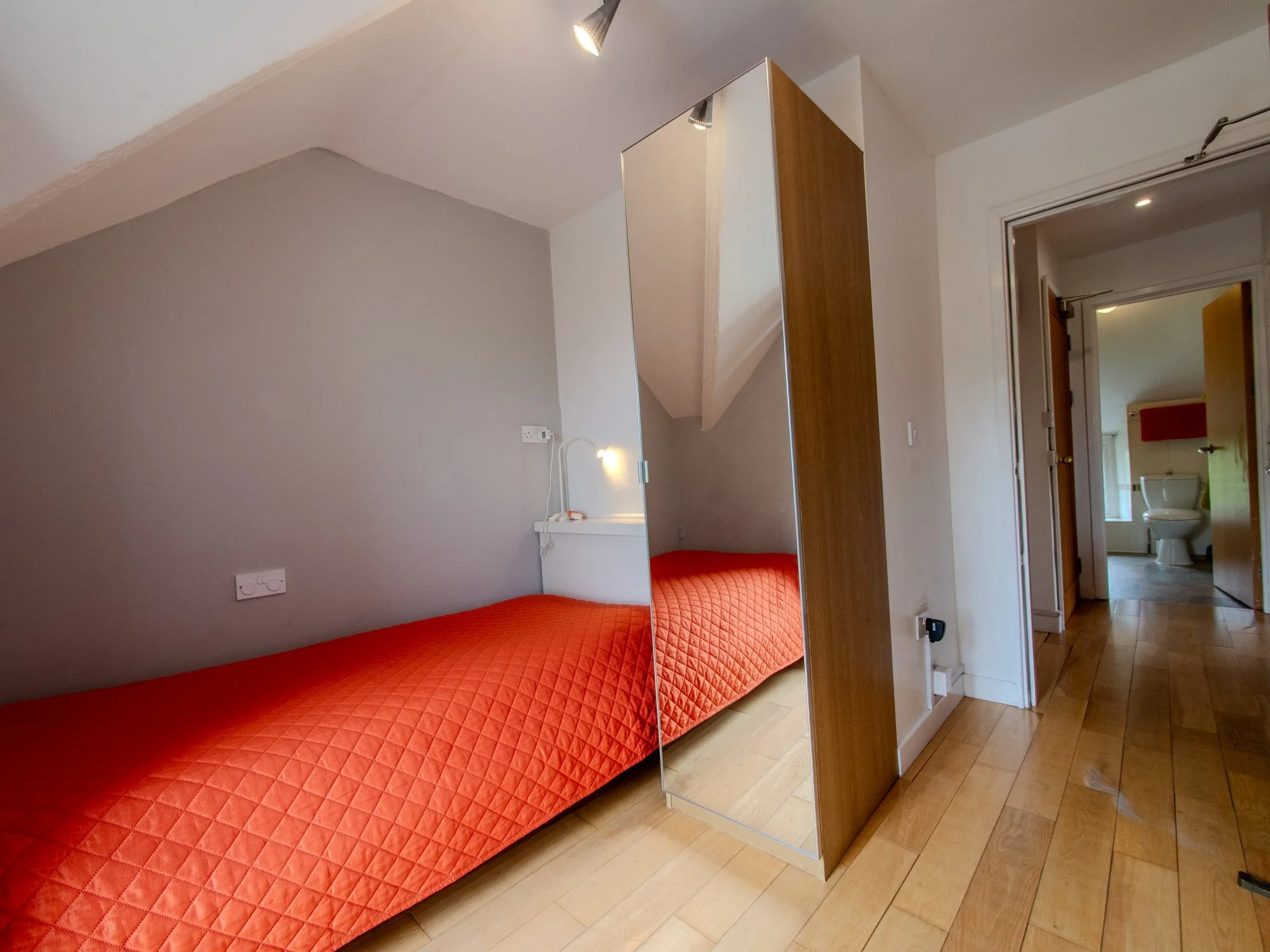 A small bedroom with a sloped ceiling, a red quilted bed with no pillows, a large mirror on a wooden partition, a wall-mounted reading light, and an open door leading to a bathroom with a toilet visible.
