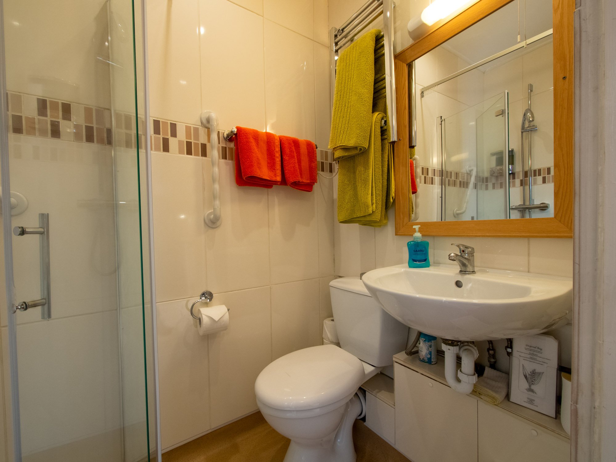 A small bathroom with a shower stall, a mirror with a wooden frame, a sink, and a toilet. Towel bars with orange and green towels and a bottle of soap on the counter.