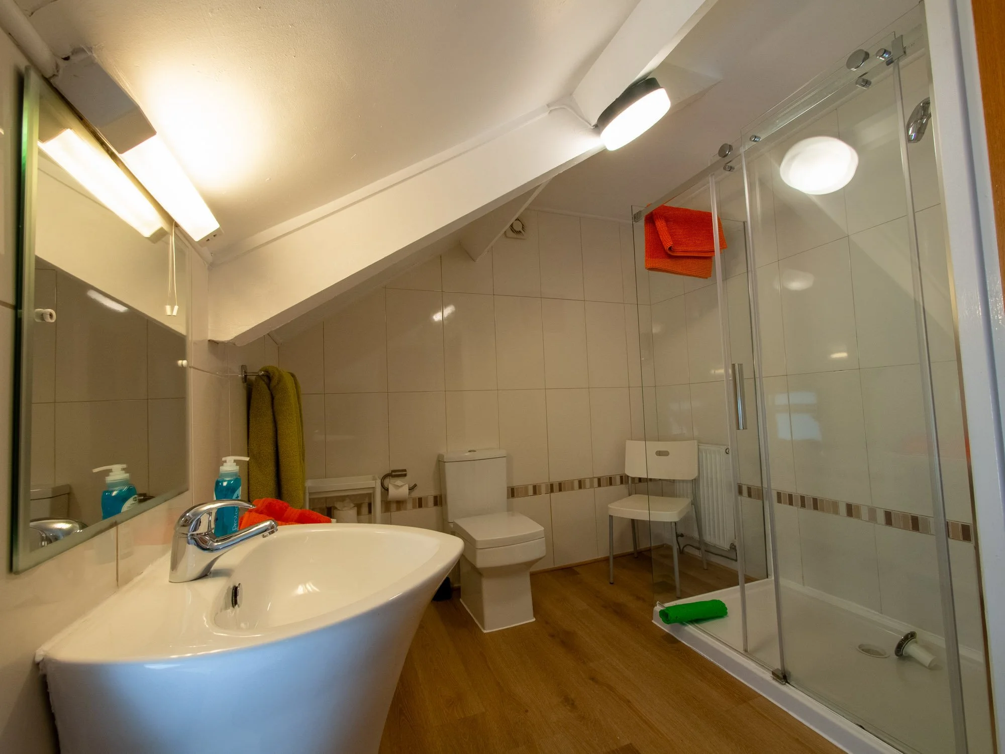 A bathroom with a white bathtub, a sink with a mirror, a toilet, a glass shower enclosure, and orange towels, with wood flooring and tiled walls.
