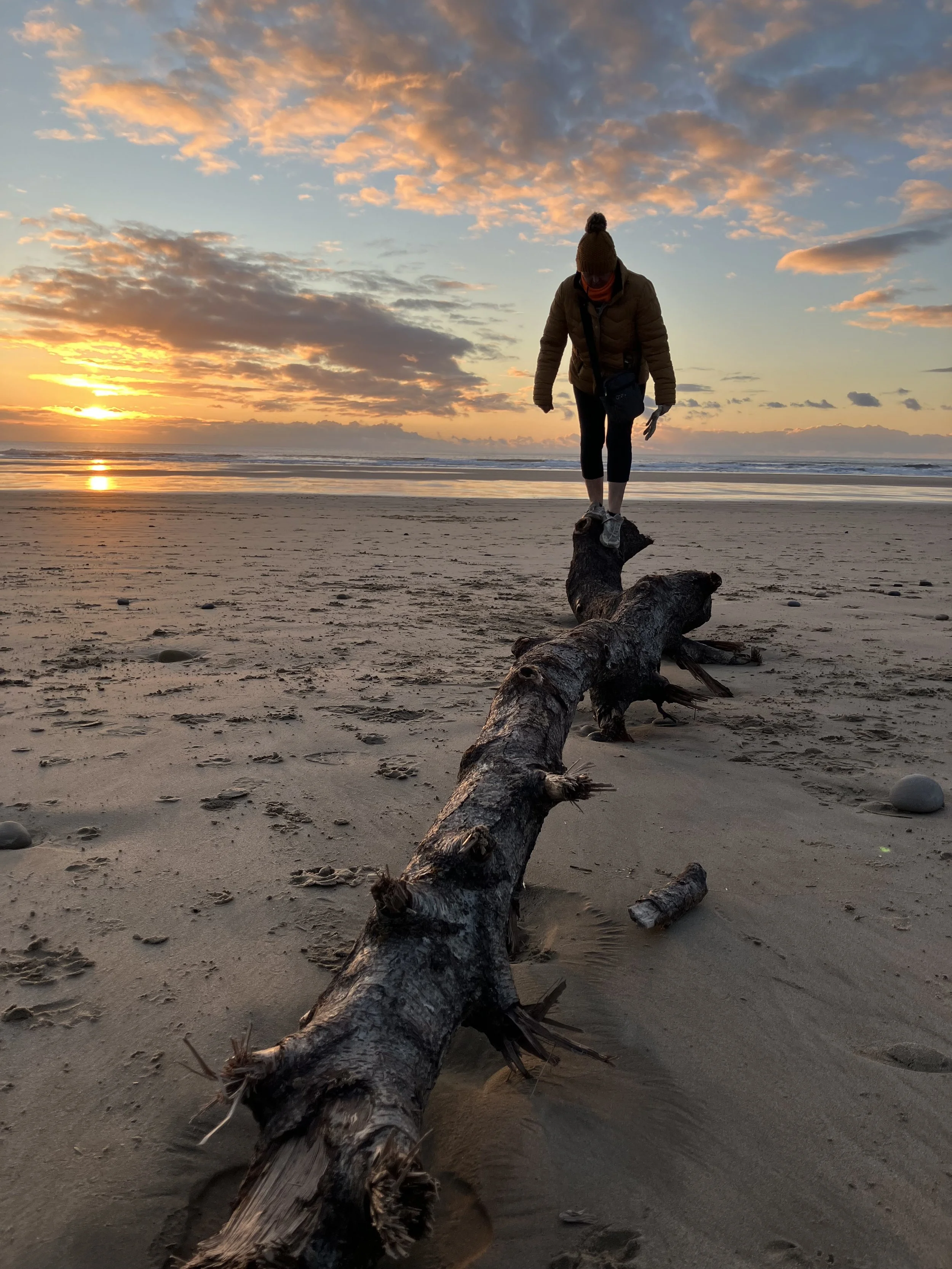 Person balancing on a driftwood log at the beach during sunset with a colorful sky in the background.