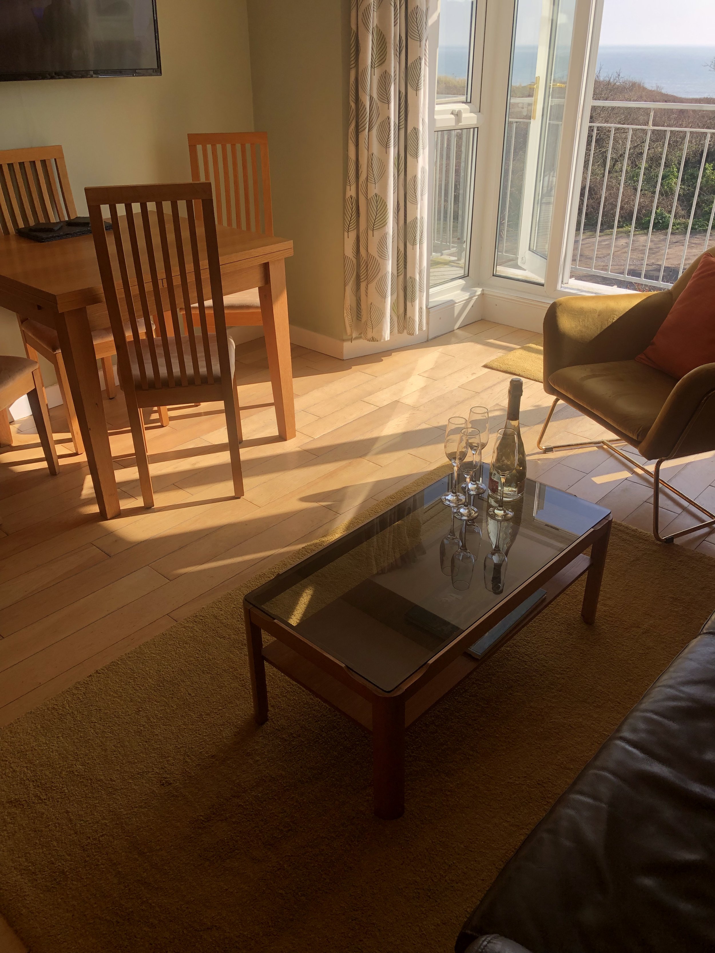Living room with a glass coffee table, three champagne flutes, a bottle, wooden dining table with chairs, and glass doors leading to a balcony with an ocean view.