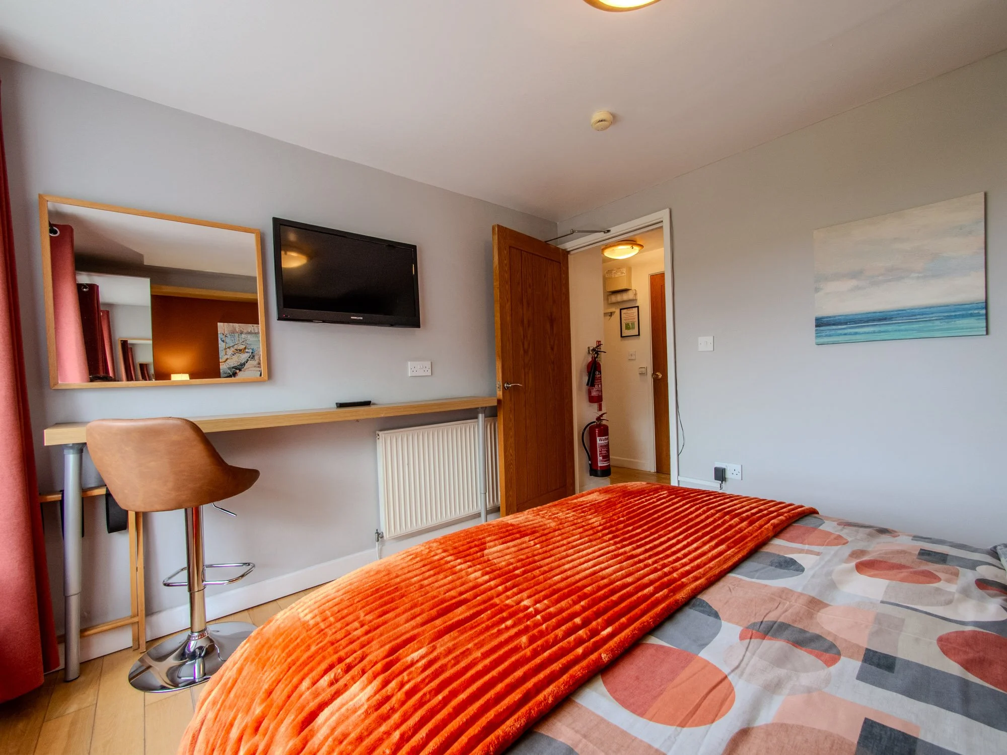A hotel room with a bed covered in a patterned comforter and an orange textured blanket, a wooden doorway, a wall-mounted TV, a mirror reflecting a framed picture, and a fire extinguisher near the door.