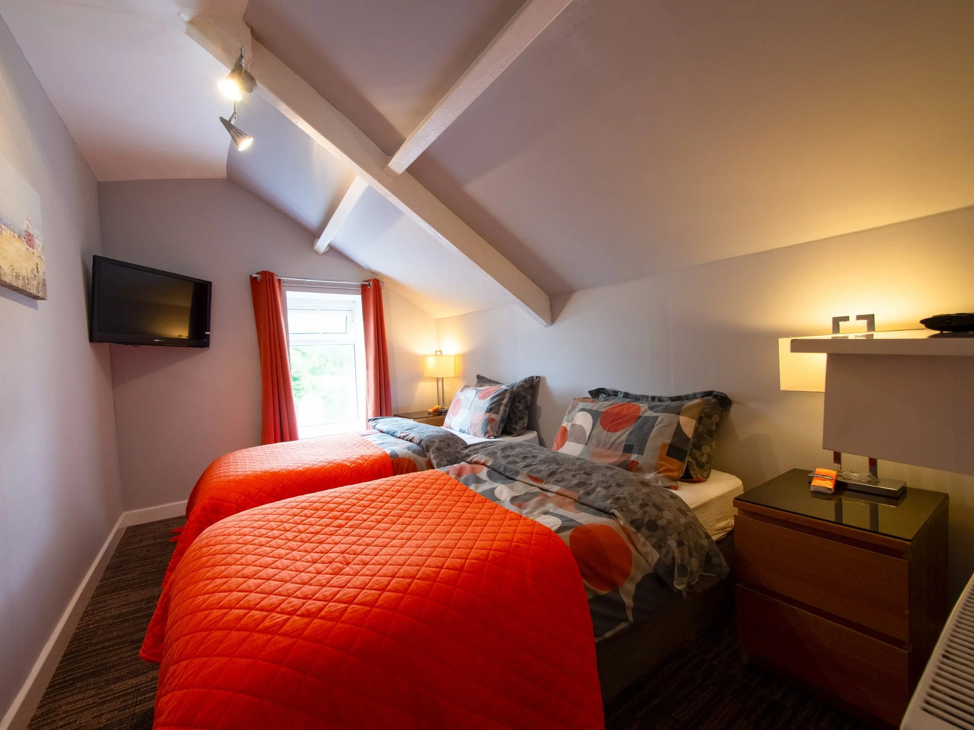 A cozy bedroom with two beds, one with a quilted orange cover and the other with a patterned gray and orange cover. There is a wall-mounted flat-screen TV, orange curtains over a window, a bedside table with a lamp, and a small shelf with a black bow