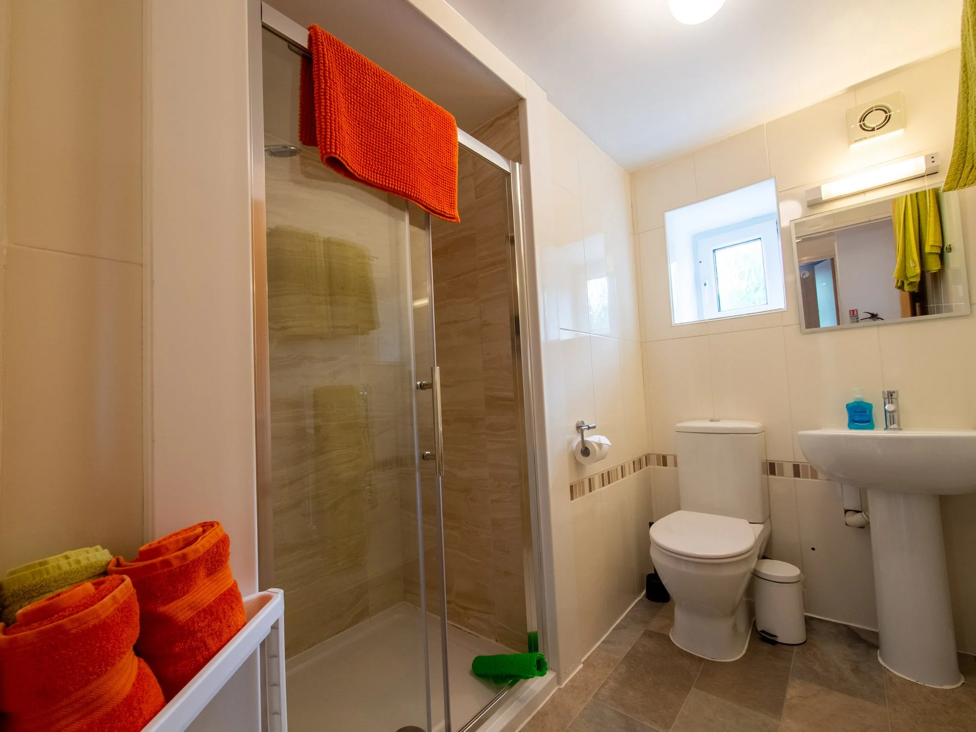 A bathroom with a shower, toilet, sink, mirror, window, and towels in orange and yellow. The shower has a glass door and beige tiles. The towels are on a shelf and hanging on hooks.
