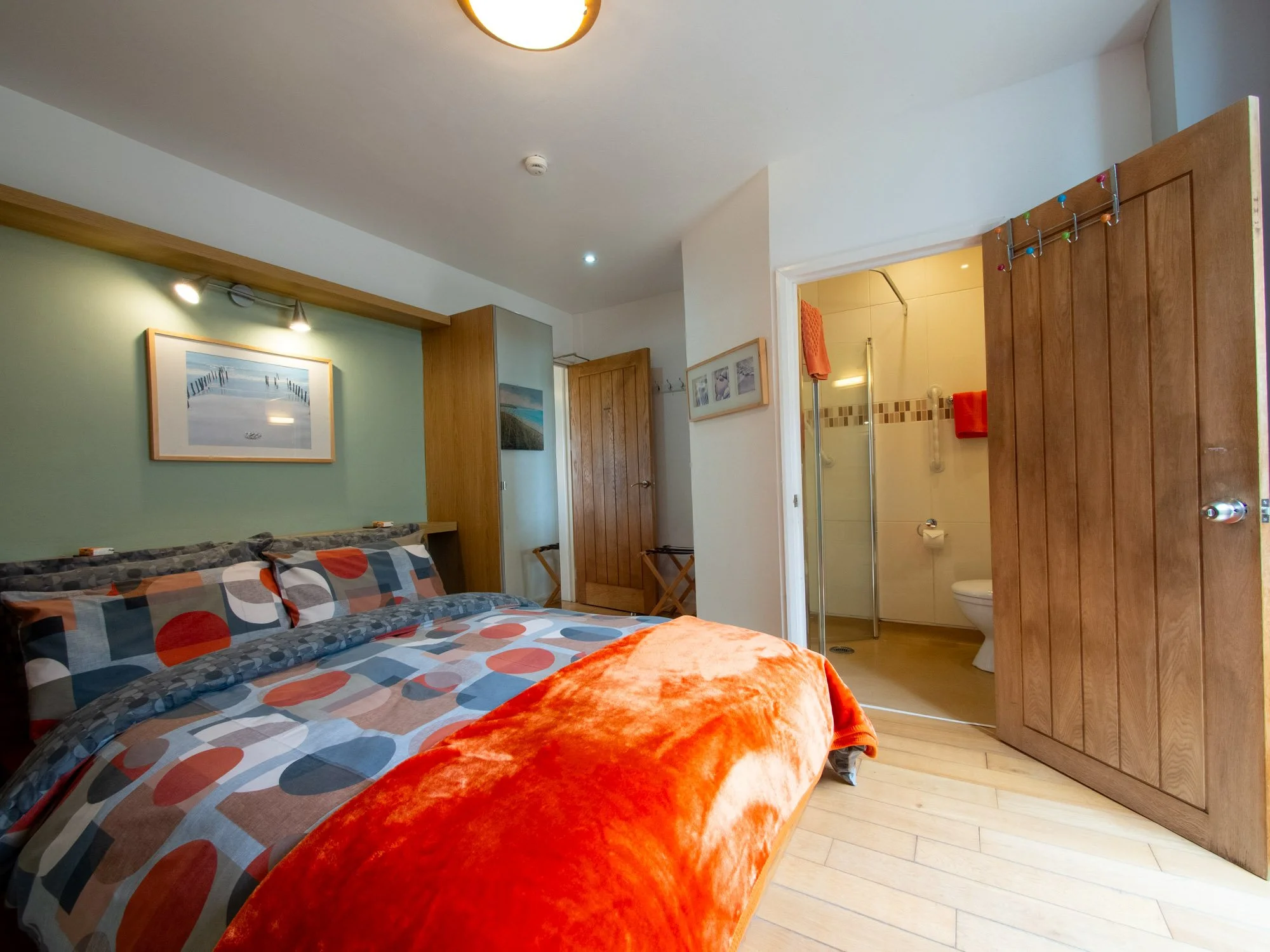 A bedroom with a colorful bedspread featuring circles in shades of orange, gray, and blue. An orange throw blanket is on the bed. There is framed artwork on a green wall above the bed, and two bedside lamps. To the right, an open door reveals a bathr