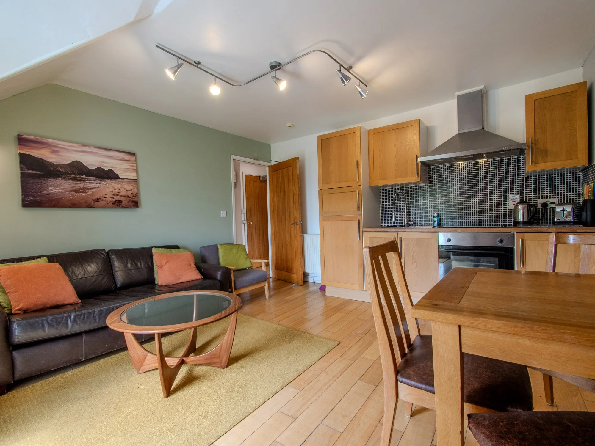 A cozy open-concept living room and kitchen area with wooden flooring, a black leather sofa with colorful cushions, a glass coffee table, a green area rug, light wood dining table with chairs, wooden kitchen cabinets, a stainless steel oven, a range 