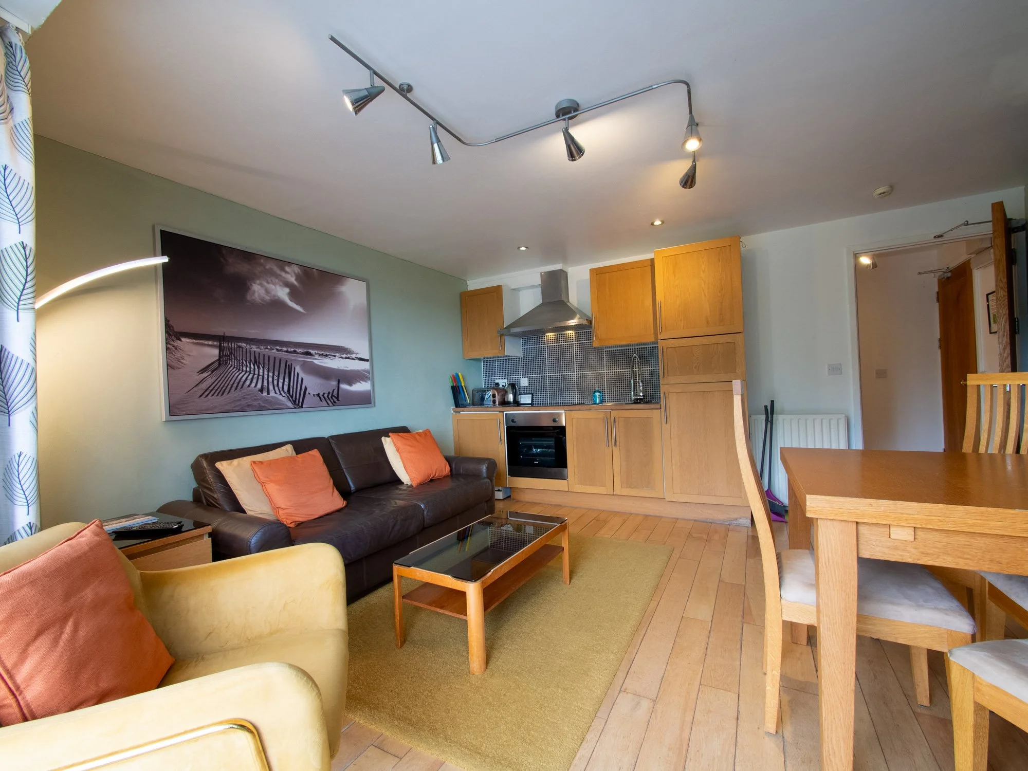 Interior of a living room with a couch, armchair, coffee table, and a kitchen in the background. There is a framed black and white beach scene on the wall, and the room has wooden flooring, a green rug, and a ceiling light fixture with multiple adjus
