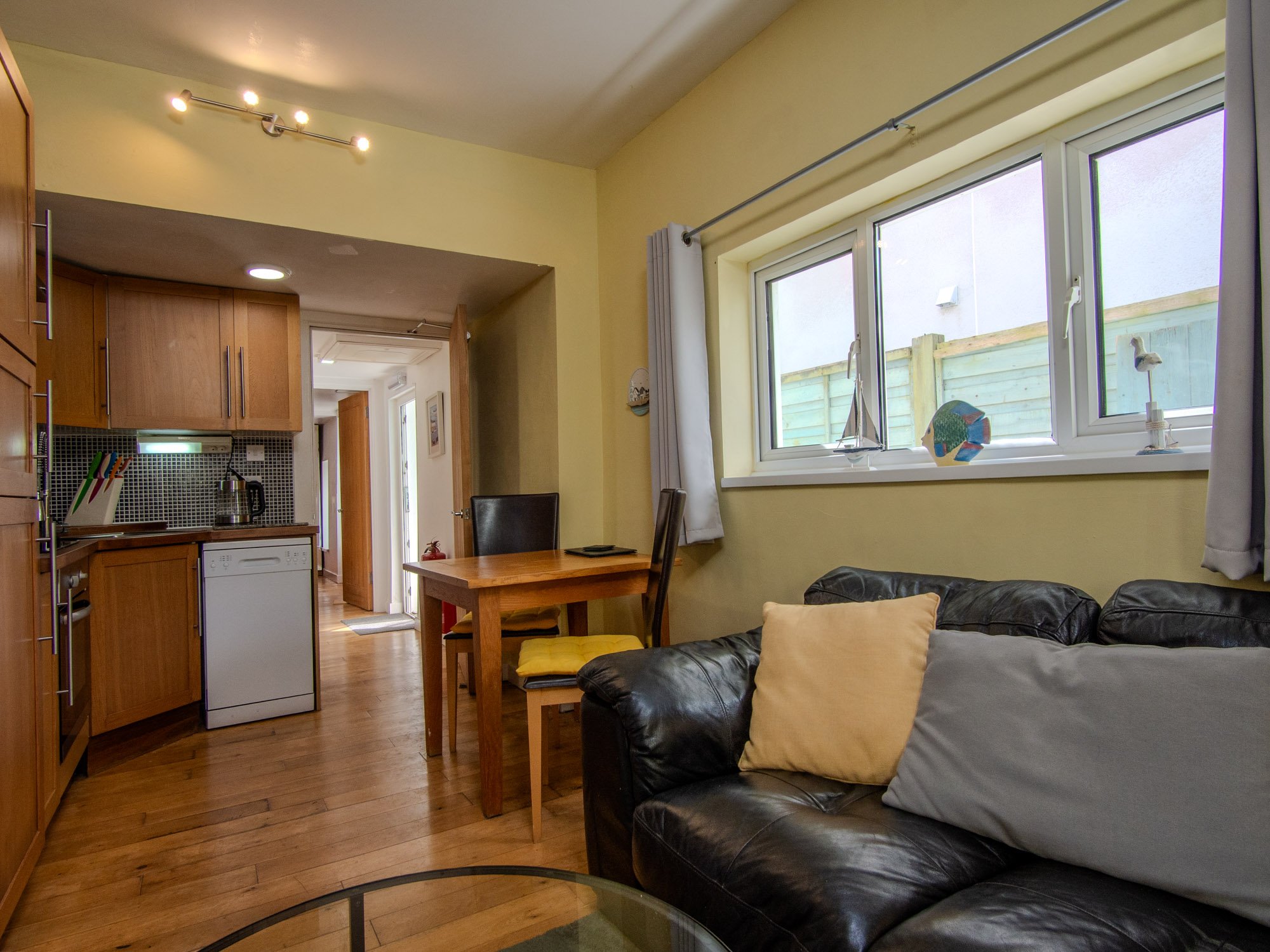 The image shows a cozy room with a black leather sofa with pillows, a wooden dining table with chairs, a kitchen area with wooden cabinets, a side door leading outside, large window with curtains, decorative items on the windowsill, and hardwood floo