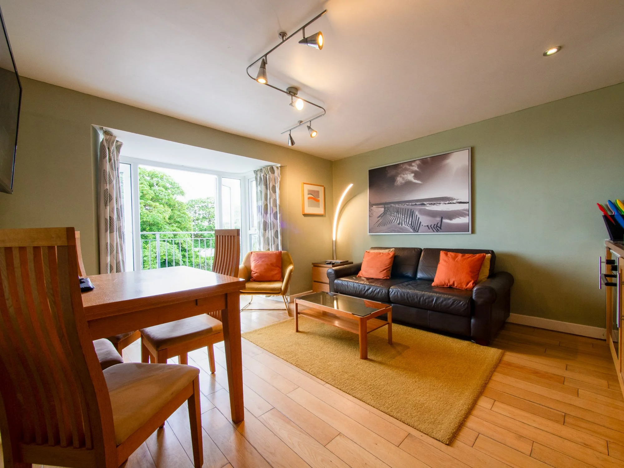Living room with wooden flooring, green walls, a black leather sofa with orange pillows, a large wall art of a beach scene, a window with curtains, a wooden dining table with chairs, and a metal floor lamp.