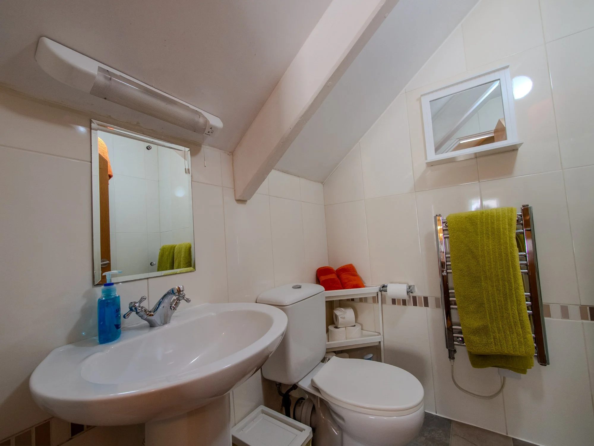 A small bathroom with a white sink, a toilet, and a rectangular mirror above the sink. A window with partial view of the outside is on the wall. Towels are hanging on a heated towel rail, with one green and others orange. There is a small white stora