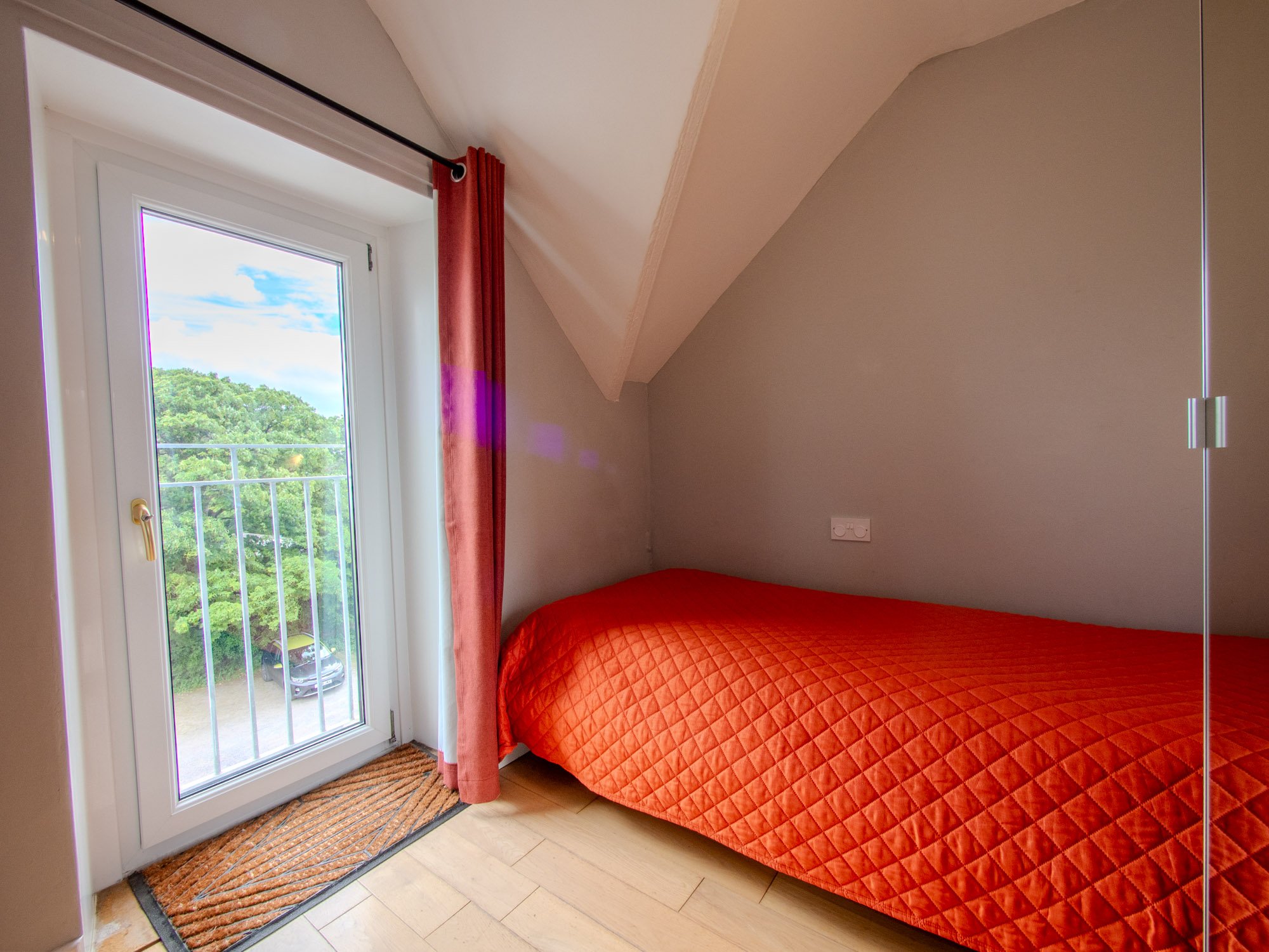 A bedroom corner with a window and a balcony door, a red quilted bedspread, an orange curtain, and a view of green trees outside.