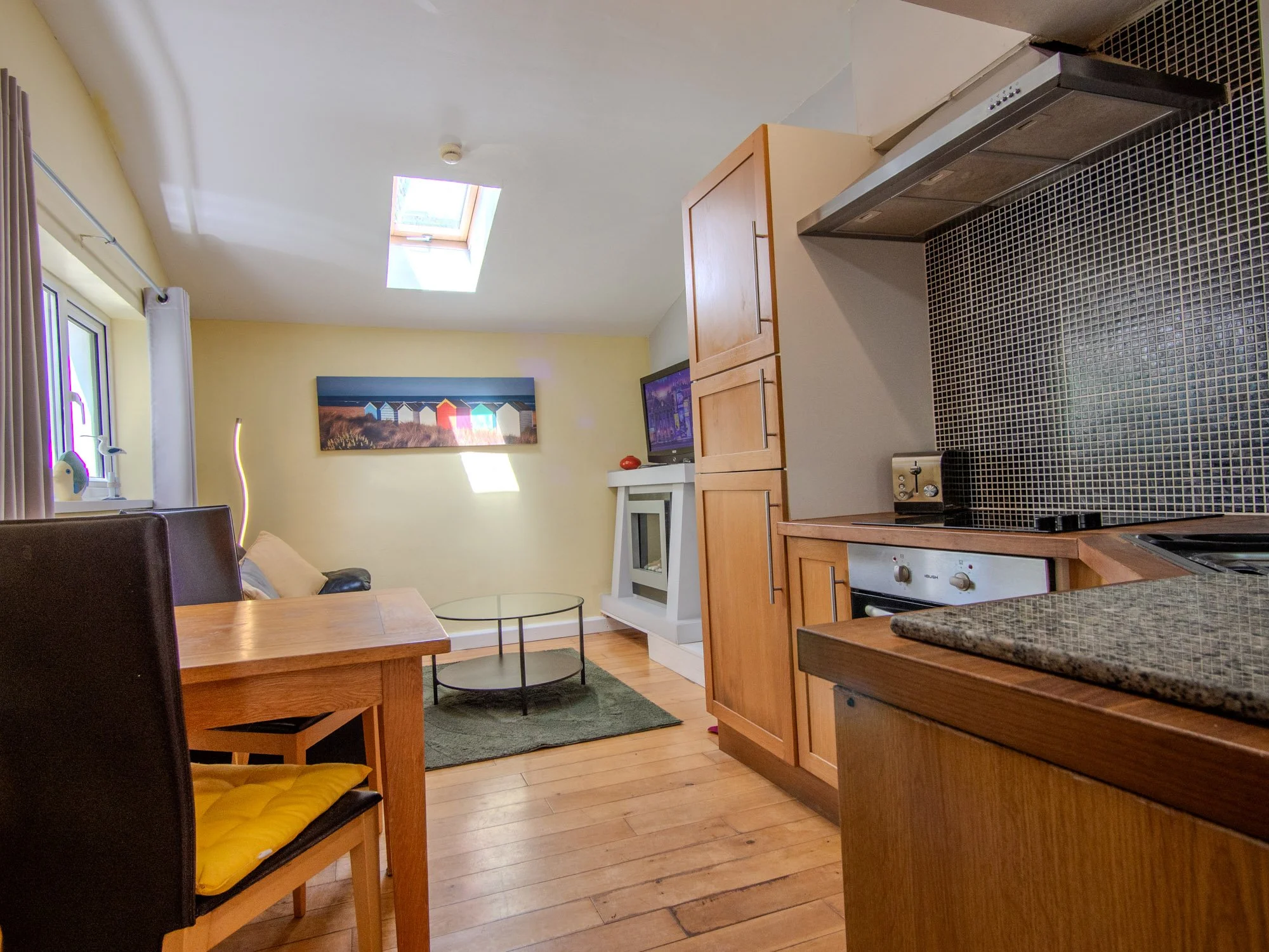 A cozy kitchen and living area with wooden flooring, a black-and-white tiled countertop, wooden cabinets, a window with curtains, a portrait painting on the wall, and a ceiling skylight allowing natural light into the room.