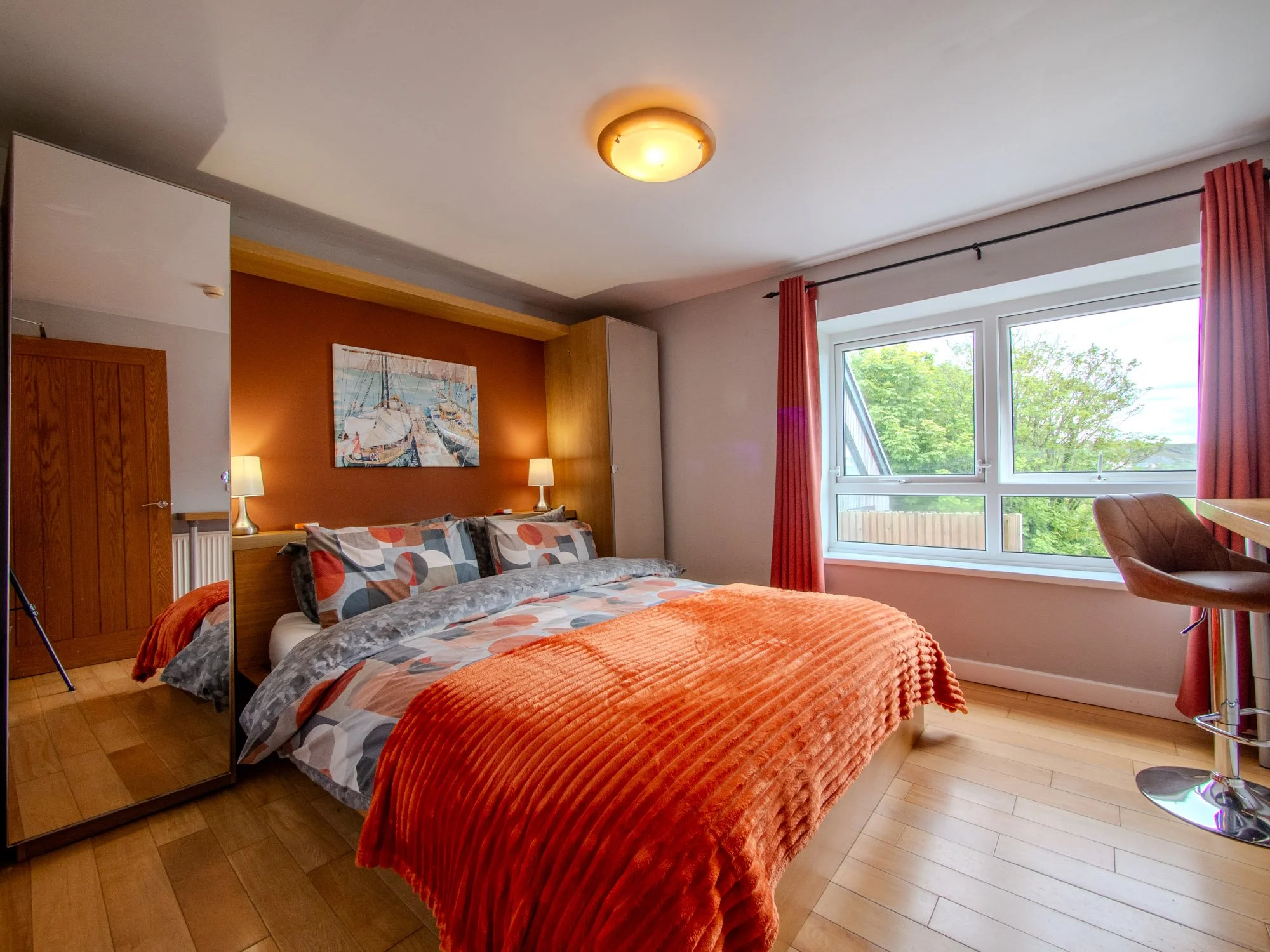 A bedroom with a bed covered in an orange textured blanket and patterned pillows, a wooden wardrobe, two lamps, a painting of boats, a large window with pink curtains, and a wooden chair.