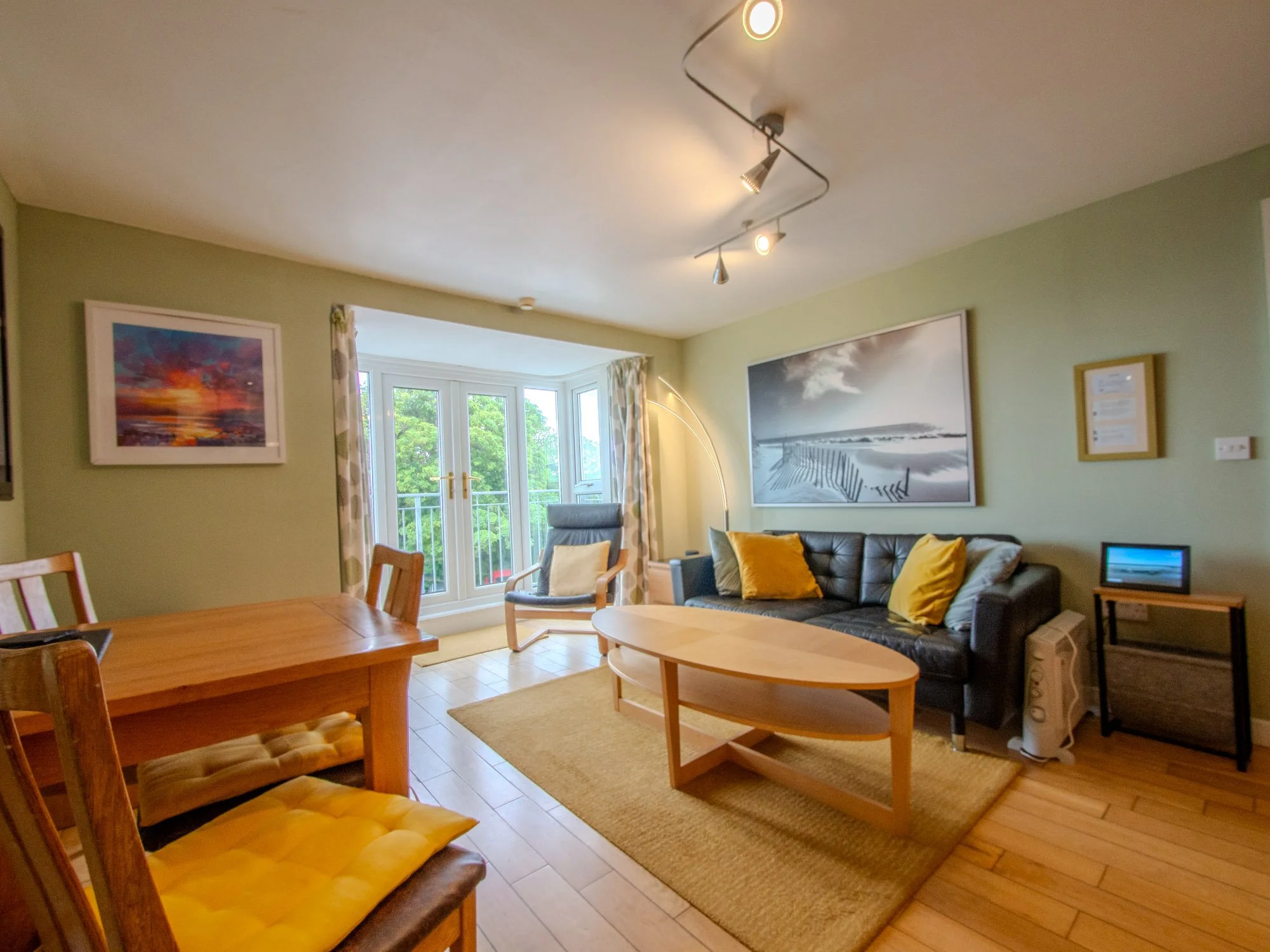 Living room with a sofa, armchair, and wooden coffee table, decorated with yellow pillows. There are large windows with curtains, and framed artwork on light green walls. A standing lamp and ceiling lighting are also visible.
