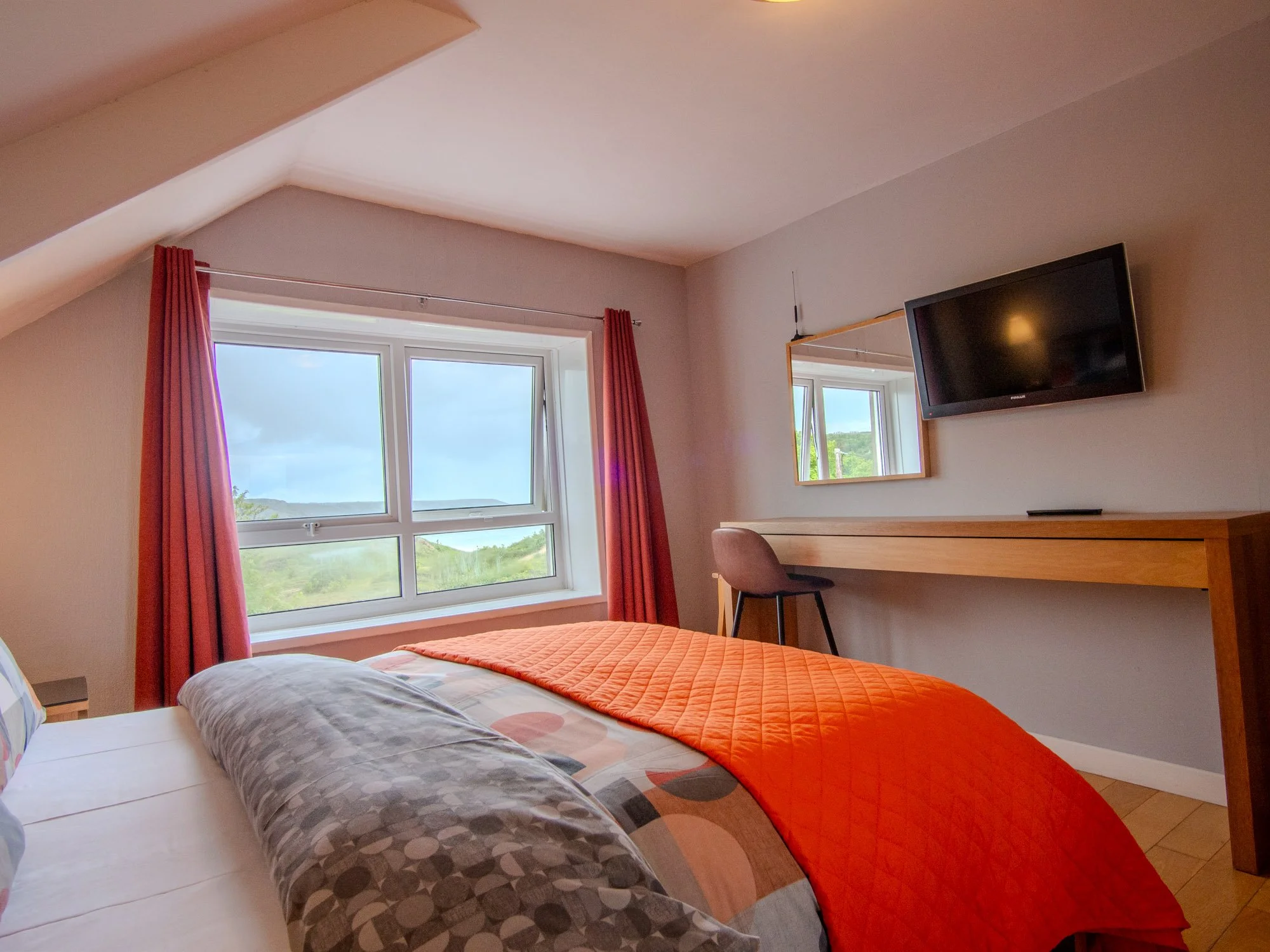 A bedroom with two beds, one with colored pillow and patterned sheets, the other with a gray pillow and patterned sheets. There is a window with pink curtains, a wall-mounted TV, and a mirror on the wall. The room has a sloped ceiling and a view of g
