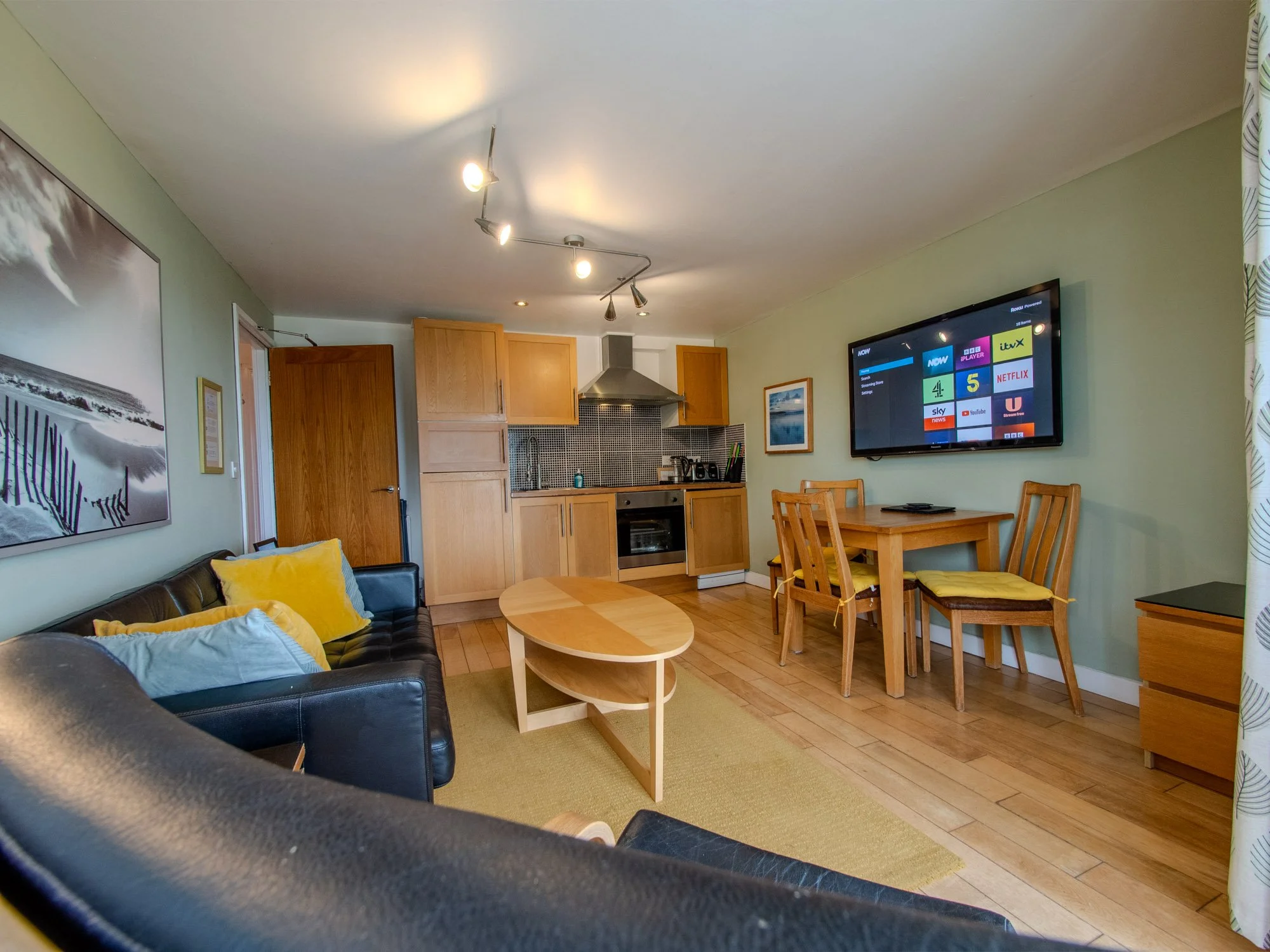 Living room with black leather sofa, yellow and light blue pillows, round wooden coffee table, green rug, kitchen with wooden cabinets, stainless steel oven, wall-mounted TV displaying streaming service icons, dining table with four chairs, window wi