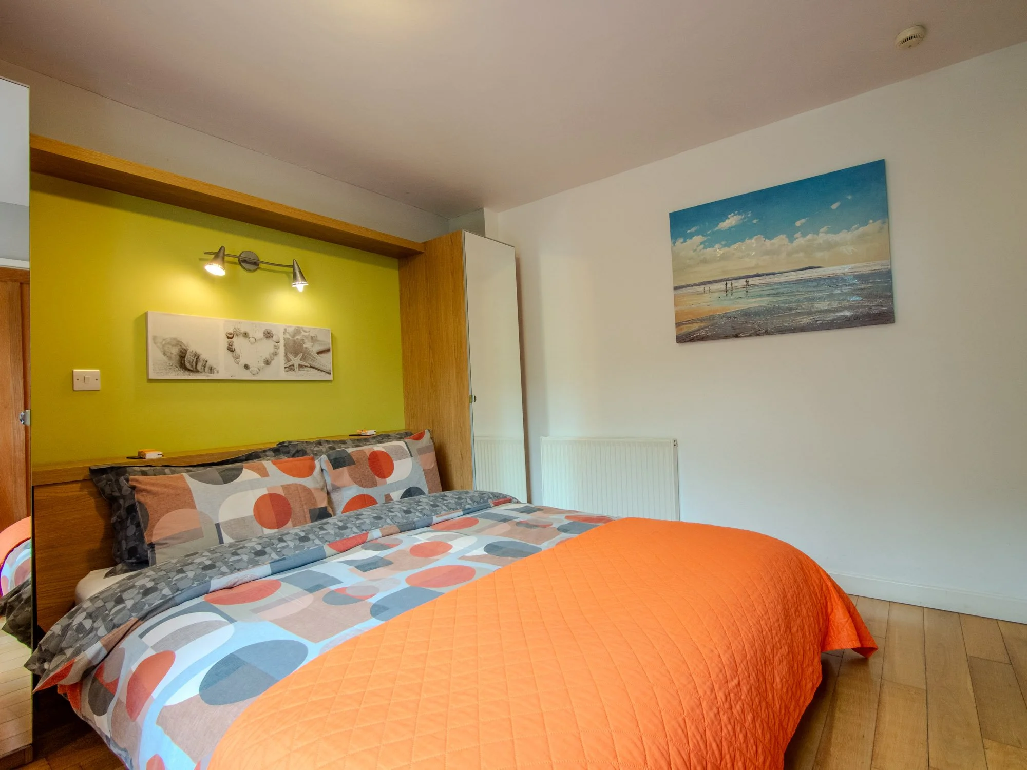 A bedroom with a bed featuring a geometric patterned quilt and pillows, a green accent wall with wall art, and a large beach painting on the wall.