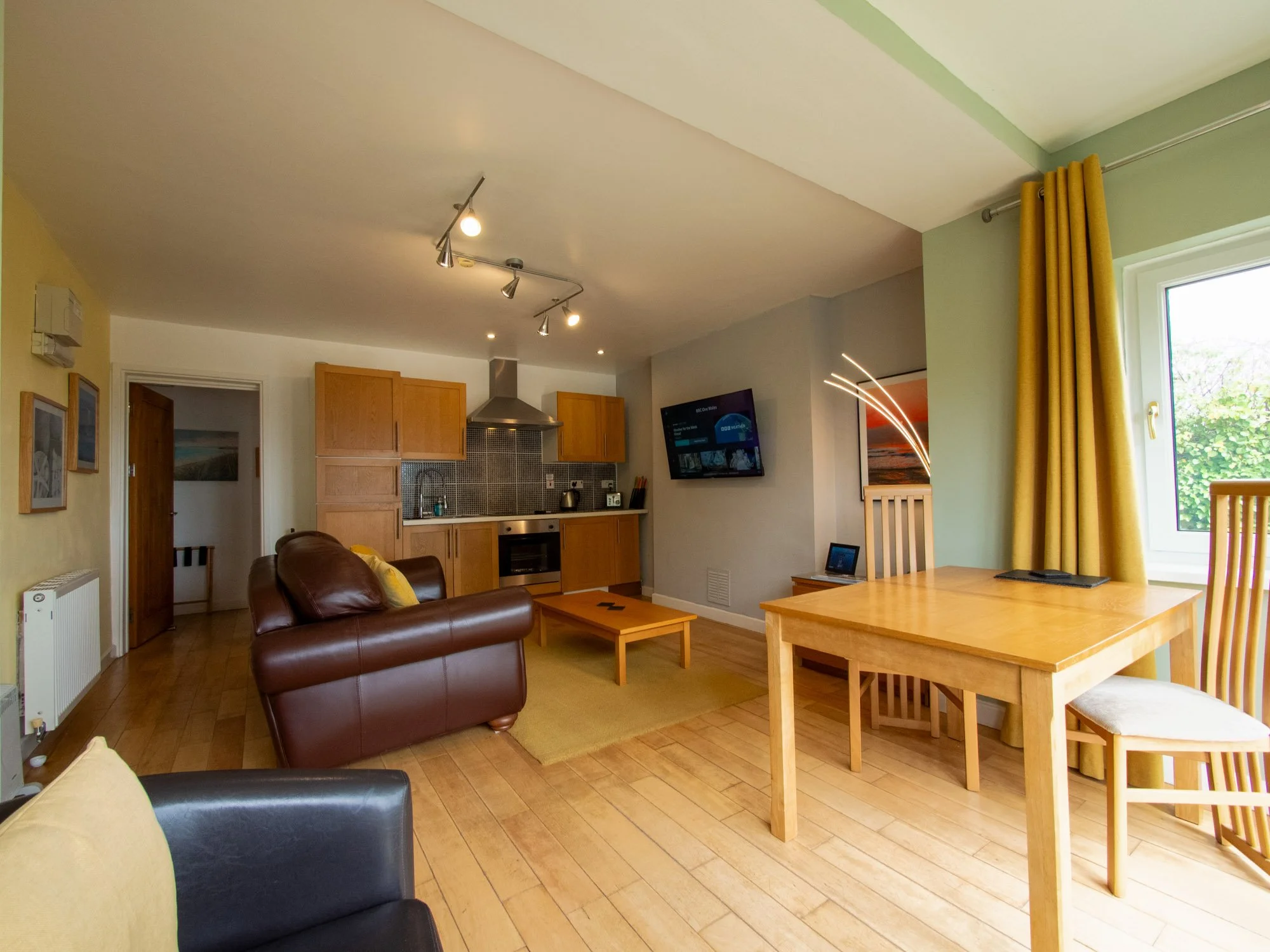 Living room with wooden flooring, yellow walls, a brown leather sofa with yellow cushions, a wooden dining table with chairs, a kitchen with wooden cabinets, a gray wall with a mounted TV, yellow curtains, and a large window showing greenery outside.