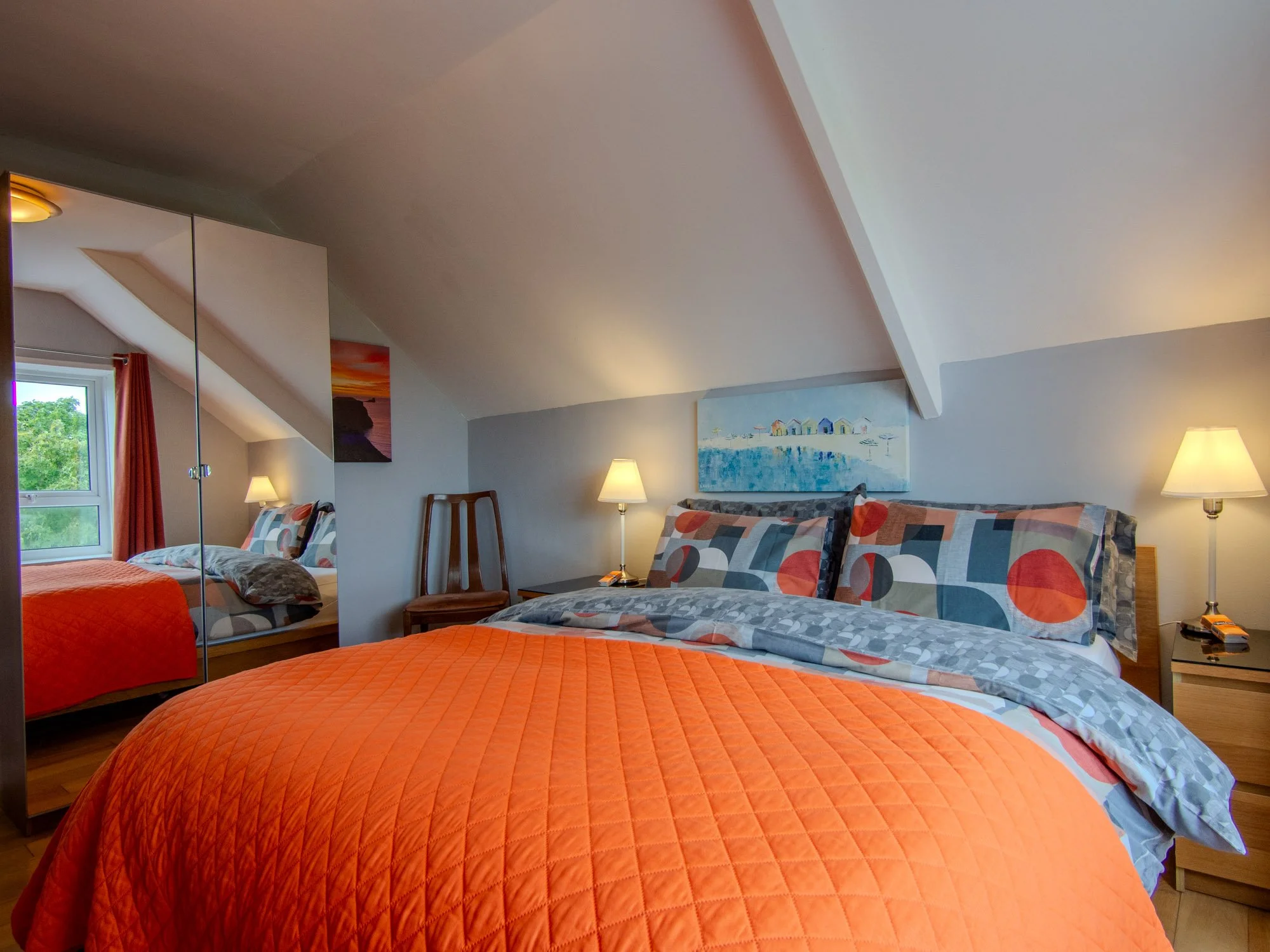 A bedroom with two beds, both with orange quilted bedspreads and decorative pillows featuring a circular pattern. There's a window with orange curtains, a mirror with sliding doors, a painting of a beach scene, and two lamps giving warm lighting. The