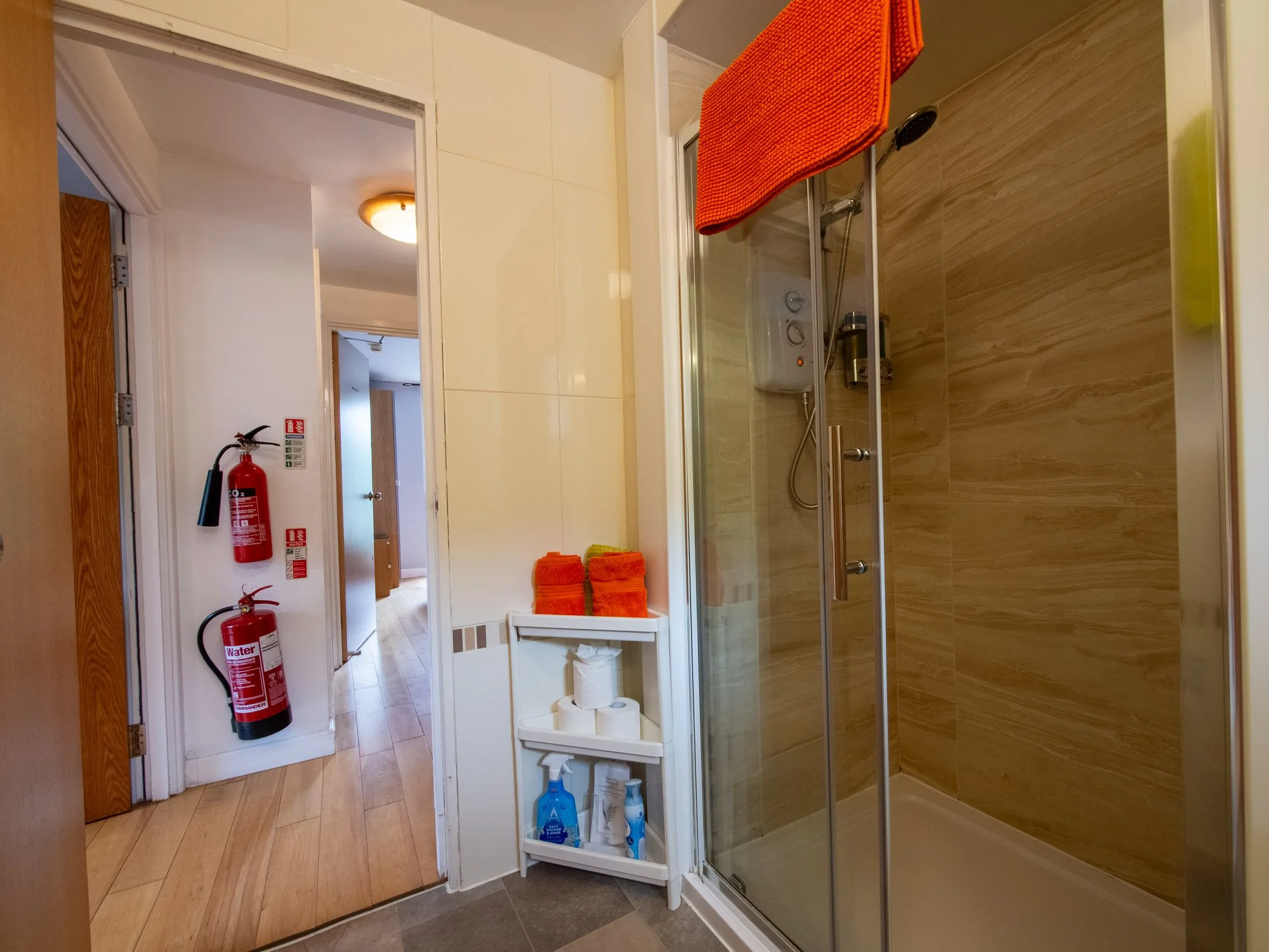 A bathroom with a glass shower stall, an orange towel hanging inside, a small white shelf with toiletries, and the hallway with wooden flooring leading to bedroom doors. Fire extinguishers are mounted on the wall near the door.