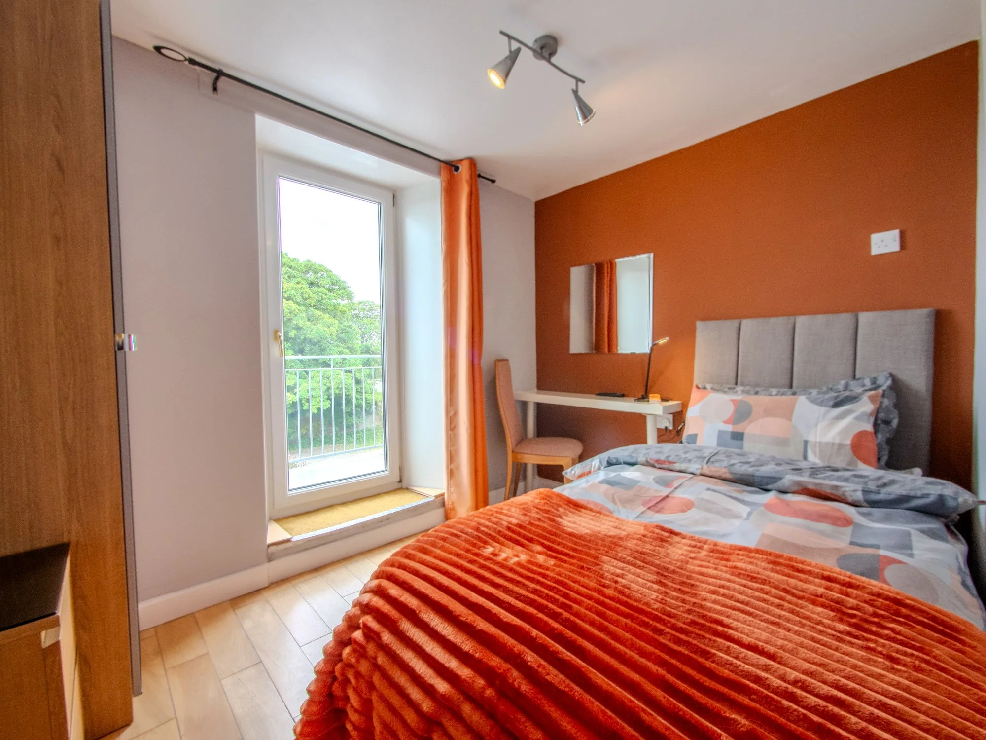 A bedroom with an orange accent wall, a bed with multicolored patterned bedding, a white desk with a lamp, a pink chair, a mirror on the wall, and a door leading to a balcony with a view of green trees.