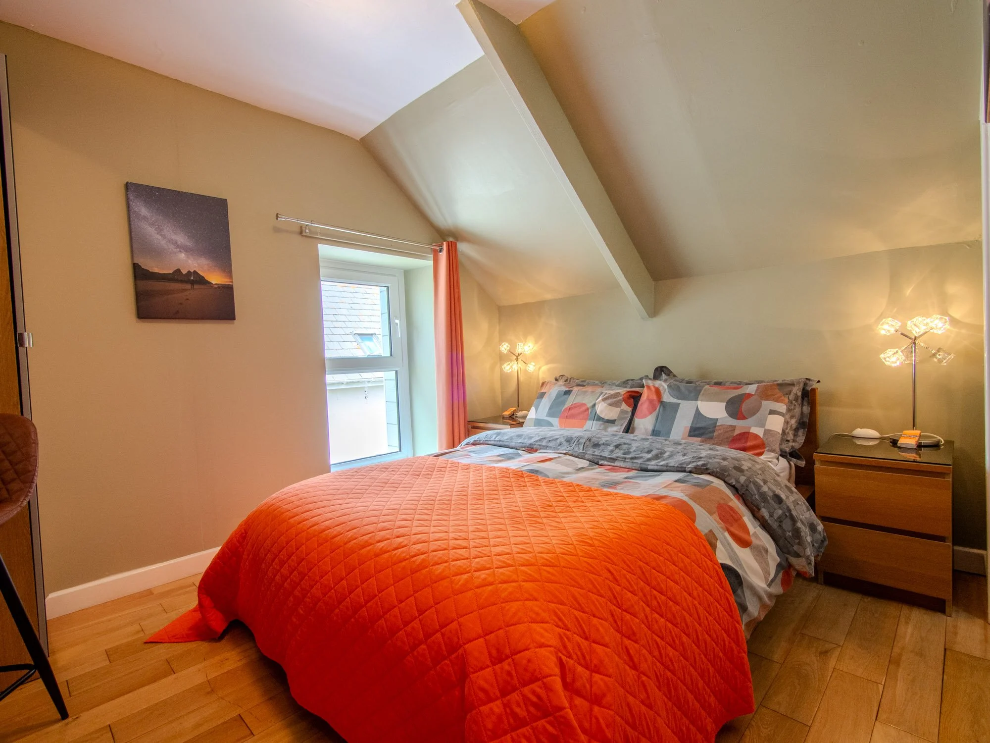 A cozy bedroom with a bed covered in a quilted orange comforter, situated near a window with beige curtains. The room has light-colored walls, a ceiling with an angled ceiling beam, and a wooden nightstand with a lamp and other small items. There is 