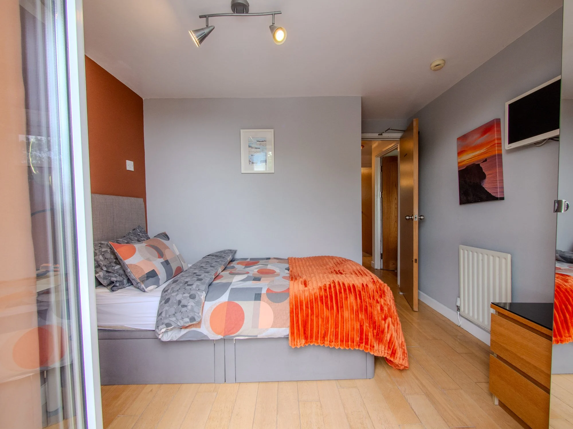 A bedroom with a double bed covered with patterned bedding, gray headboard, and an orange blanket. The room has light gray walls, a painting, a wall-mounted television, and wooden flooring. There's a wooden door, a radiator, and artwork on the wall.