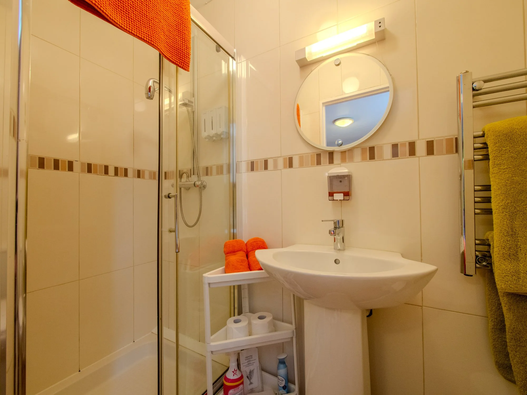 A small bathroom with beige tiled walls and a mirror above the sink. There is a glass shower enclosure with a showerhead and soap dispenser. An orange towel hangs over the shower door, and a yellow towel is on a towel rack. A white table below the mi