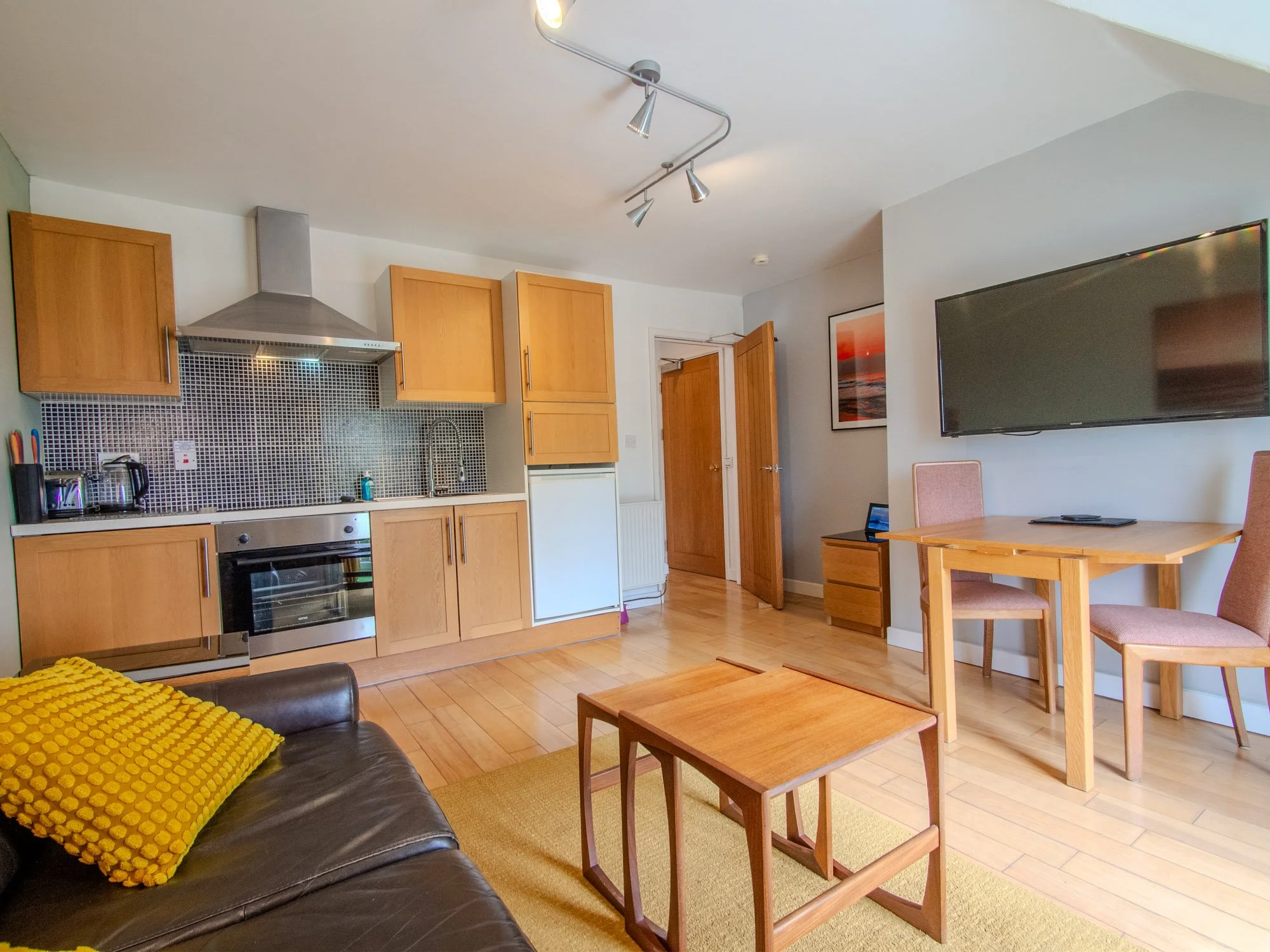 Interior view of a living room and kitchen area with wooden cabinets, a black leather sofa with a yellow throw blanket, a wooden dining table with chairs, a wall-mounted TV, and a framed ocean sunset picture.