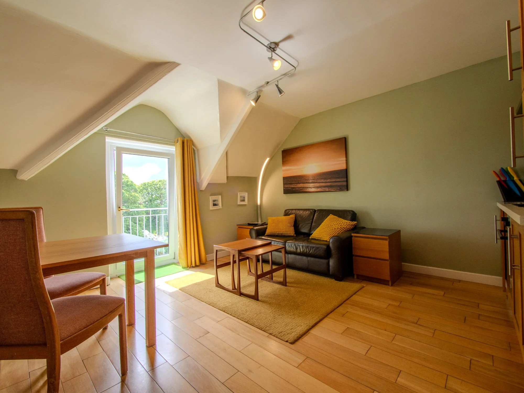 A living room with light green walls and wooden flooring, featuring a black leather sofa with yellow cushions, a wooden coffee table, a framed sunset picture on the wall, a window with yellow curtains, and ceiling lights.