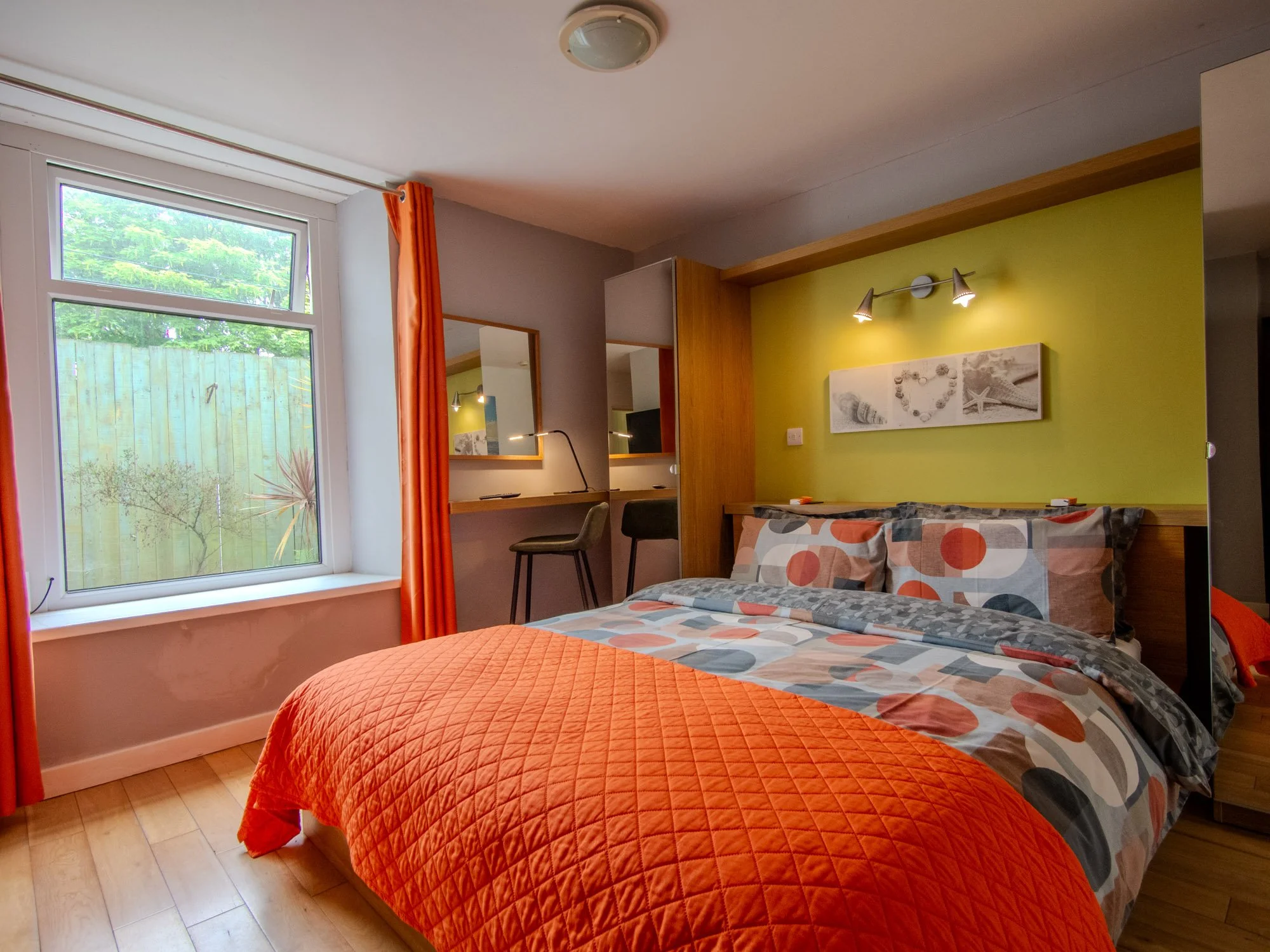 A bedroom with a large window covered by orange curtains, a bed with colorful geometric-patterned bedding and an orange quilt, a wooden headboard, a wall with a yellow accent, a modern light fixture with two spotlights, a mirror, a desk with a black 