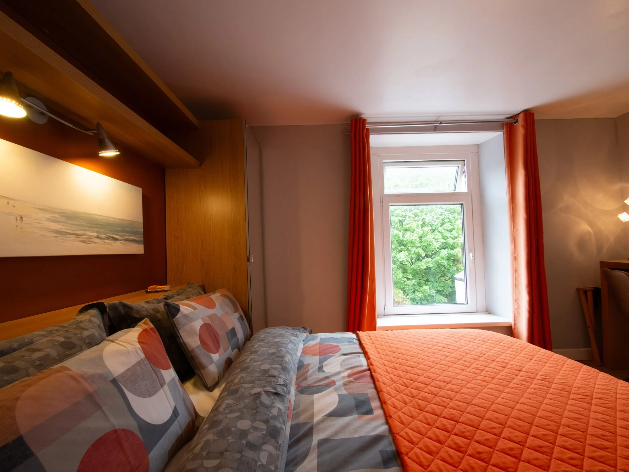 A bedroom with a window showing green trees outside, orange curtains, a bed with patterned pillows, an orange quilt, and wooden furniture.