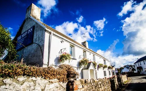 A white building with a sign reading "The Ship Inn," adorned with flower baskets, under a blue sky with clouds.