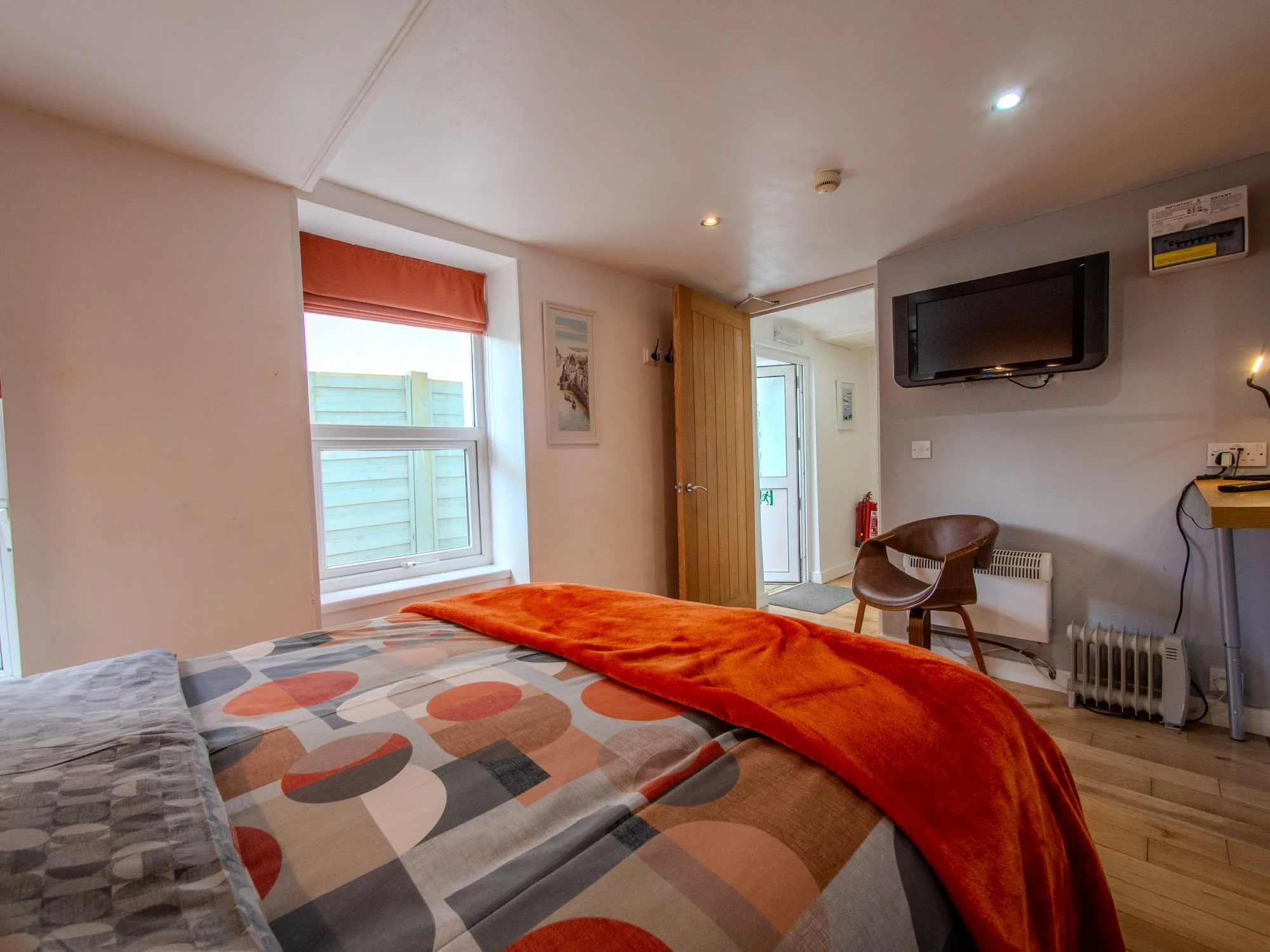 View of a cozy bedroom with a bed covered in a multi-colored polka dot duvet, an orange blanket at the foot, and pillows on top. There is a window with a red Roman shade, framed pictures on the wall, a wooden door, and a wall-mounted television. The 