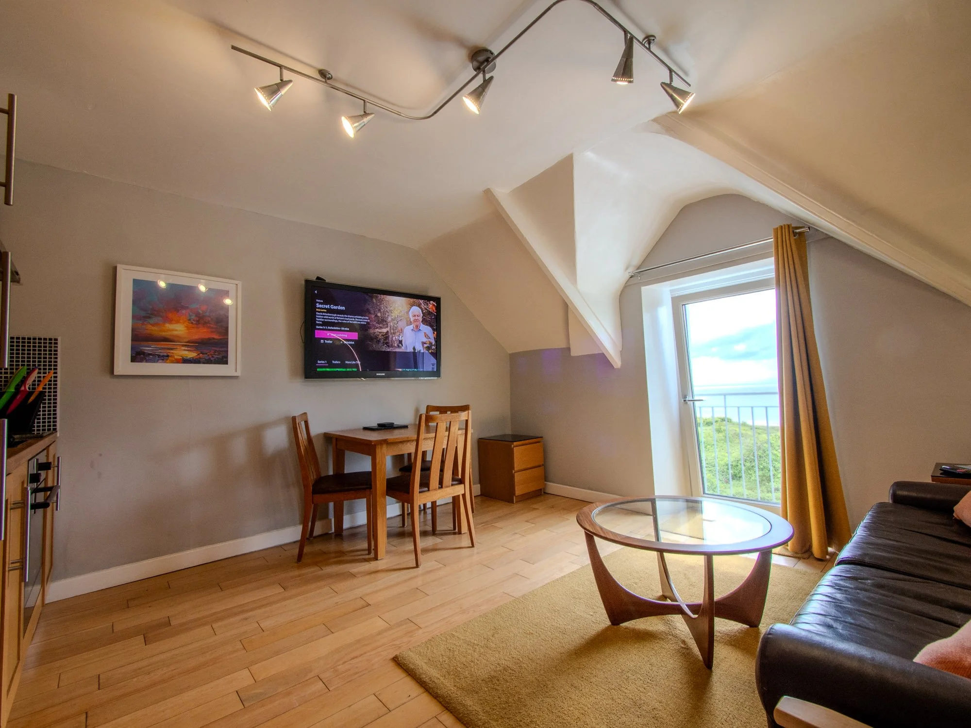 A living room with a mounted flat-screen TV, a wooden dining table with four chairs, a couch, a glass coffee table, a framed colorful sunset painting, a window with yellow curtains, and a ceiling light fixture.