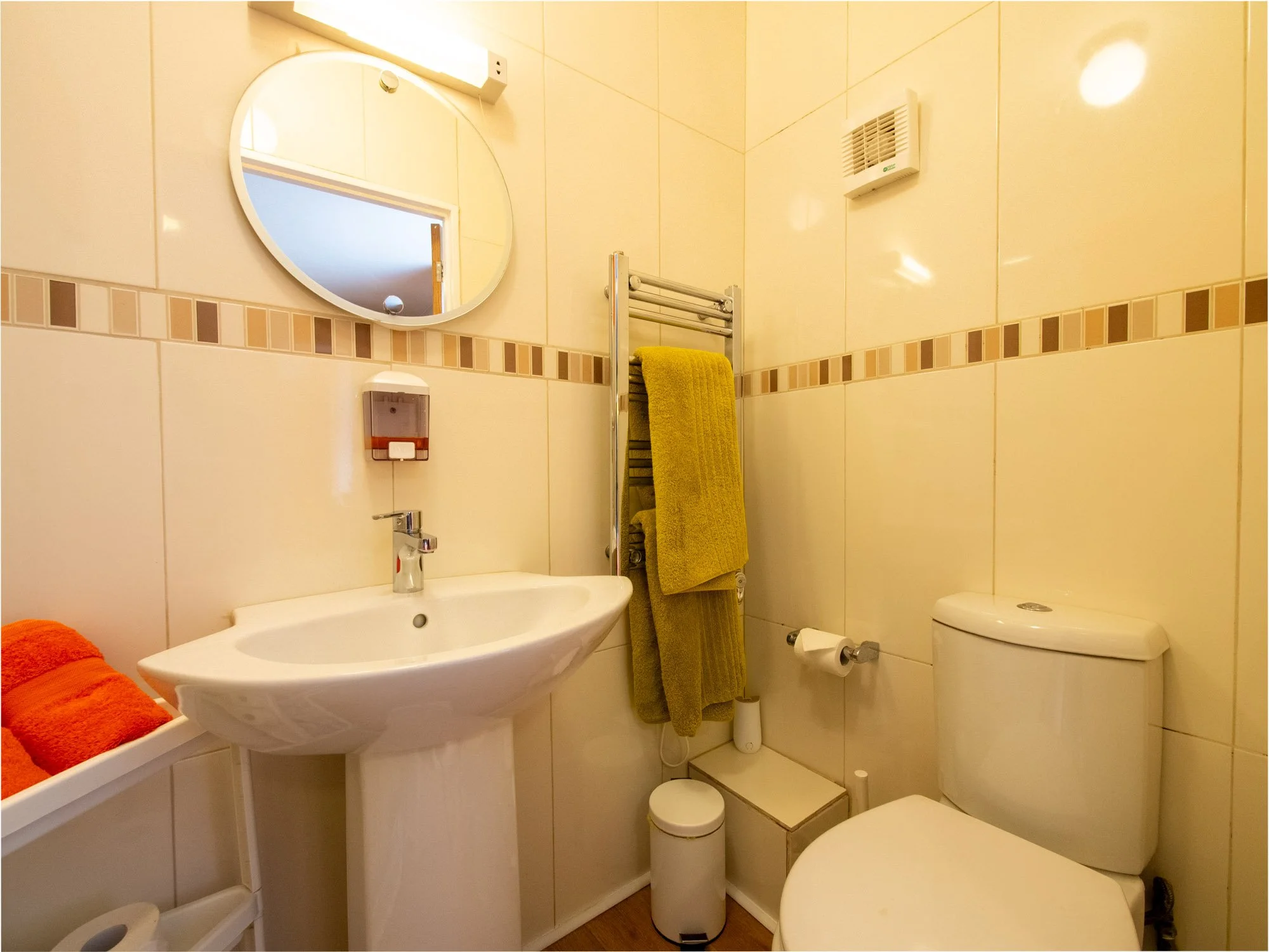 A small bathroom with beige tiled walls, a round mirror, a white sink, a yellow towel on a heated towel rail, an orange towel, a small trash bin, and a toilet. There is a vent visible on the wall and a soap dispenser above the sink.