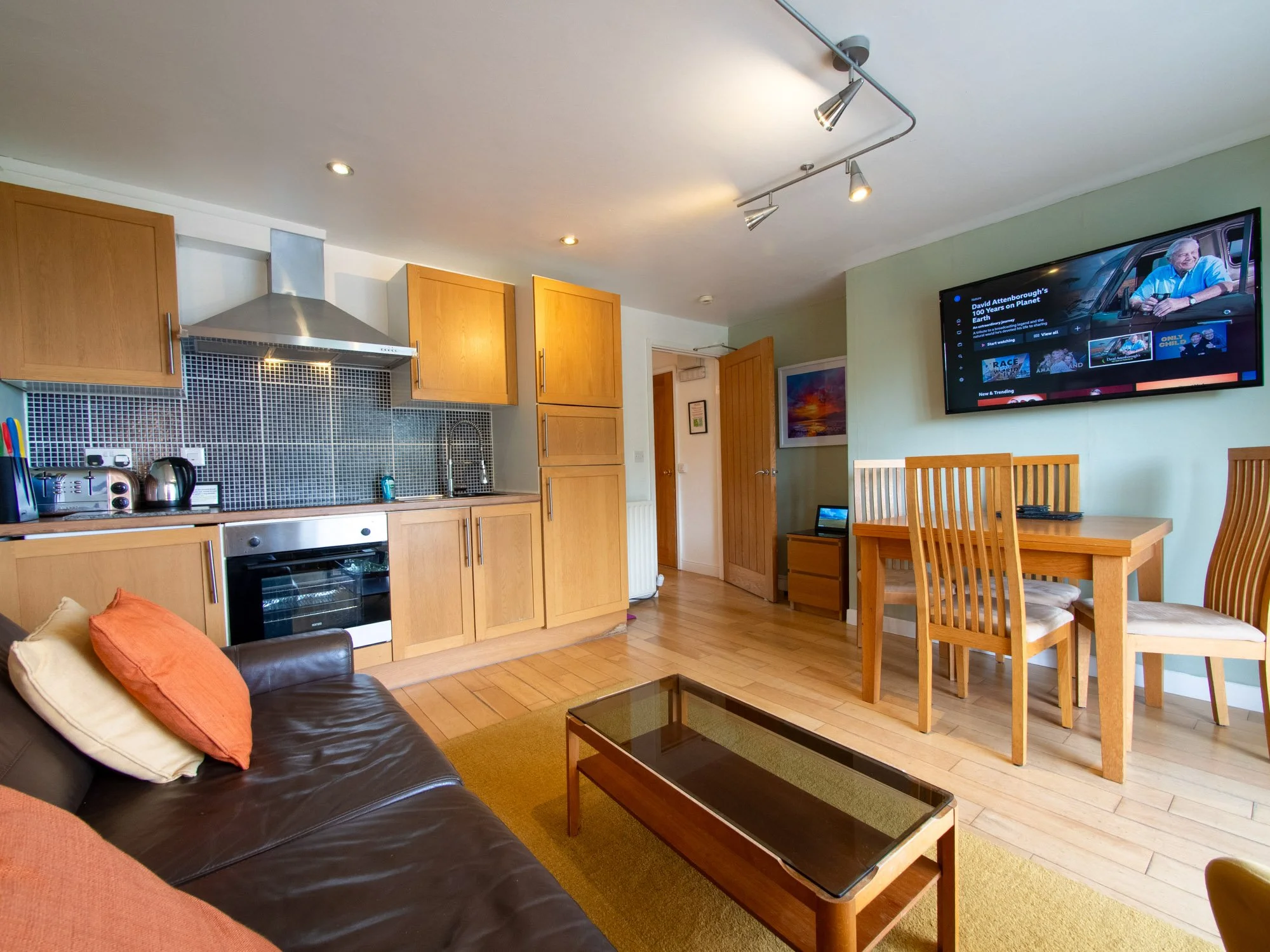 Open-plan living area with a kitchen, dining, and sitting space. There is a black leather sofa with multi-colored cushions, a glass coffee table, and a wooden dining table with several chairs. A wall-mounted flat-screen TV shows a documentary, and th