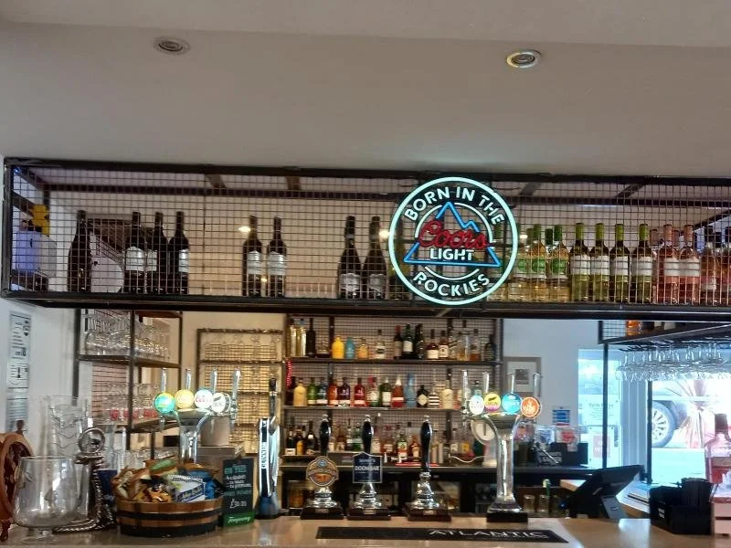 Bar counter with drink taps and a colorful assortment of bottles on shelves, including a "Born in the Rockies" Coors Light sign above.
