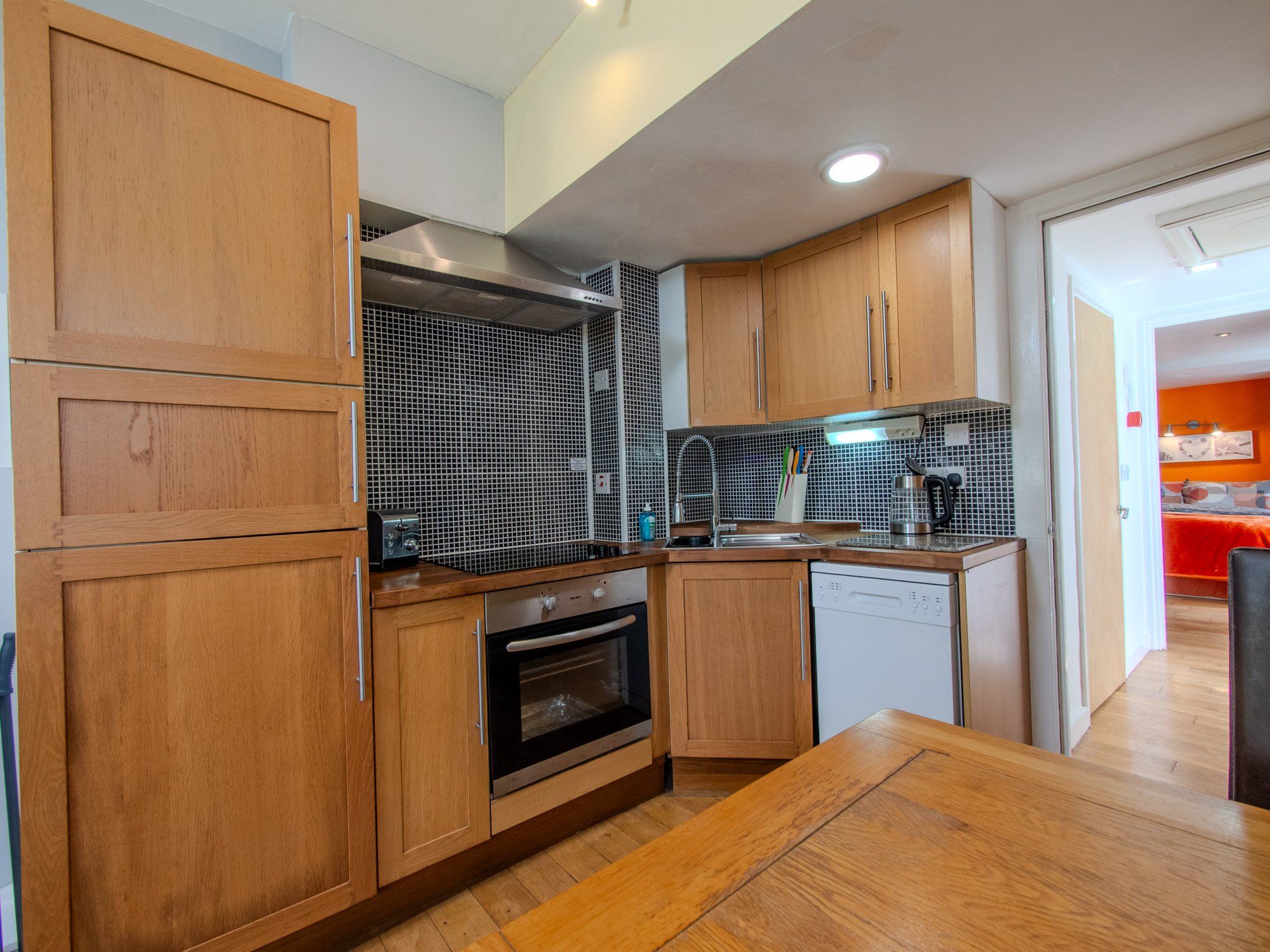 A kitchen with wooden cabinets, black mosaic tile backsplash, stainless steel oven and range hood, small countertop appliances, and a doorway leading to an orange-walled room.