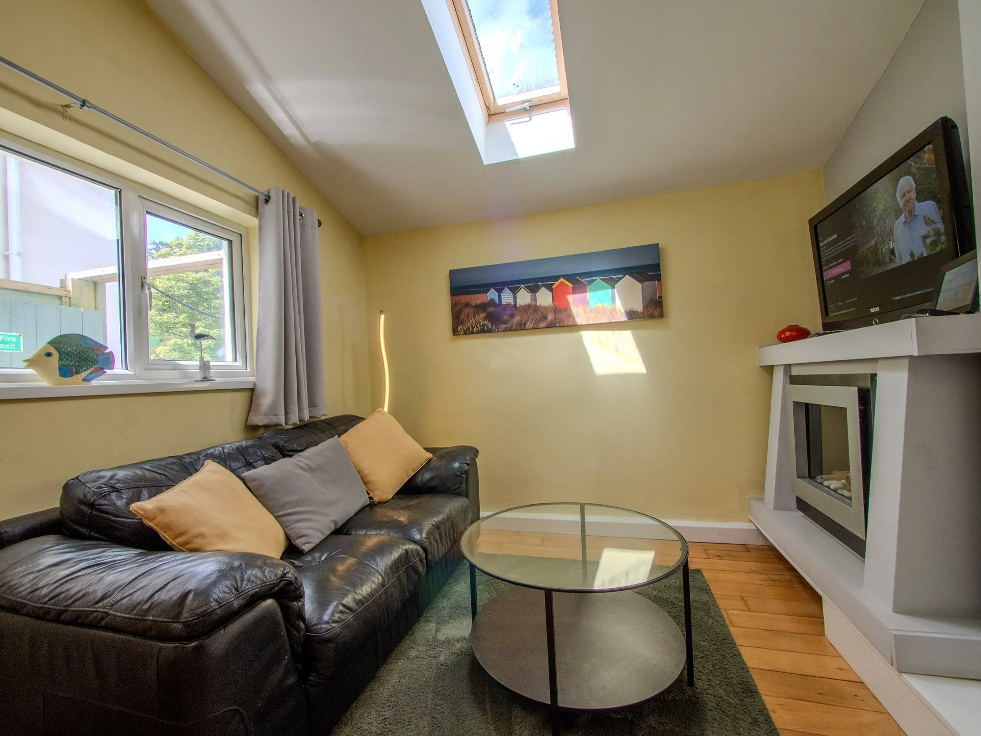 Living room with a black leather sofa, beige cushions, a round glass coffee table, yellow walls, a window with curtains, a colorful beach hut painting, a television on a white stand, a skylight, and hardwood floors.