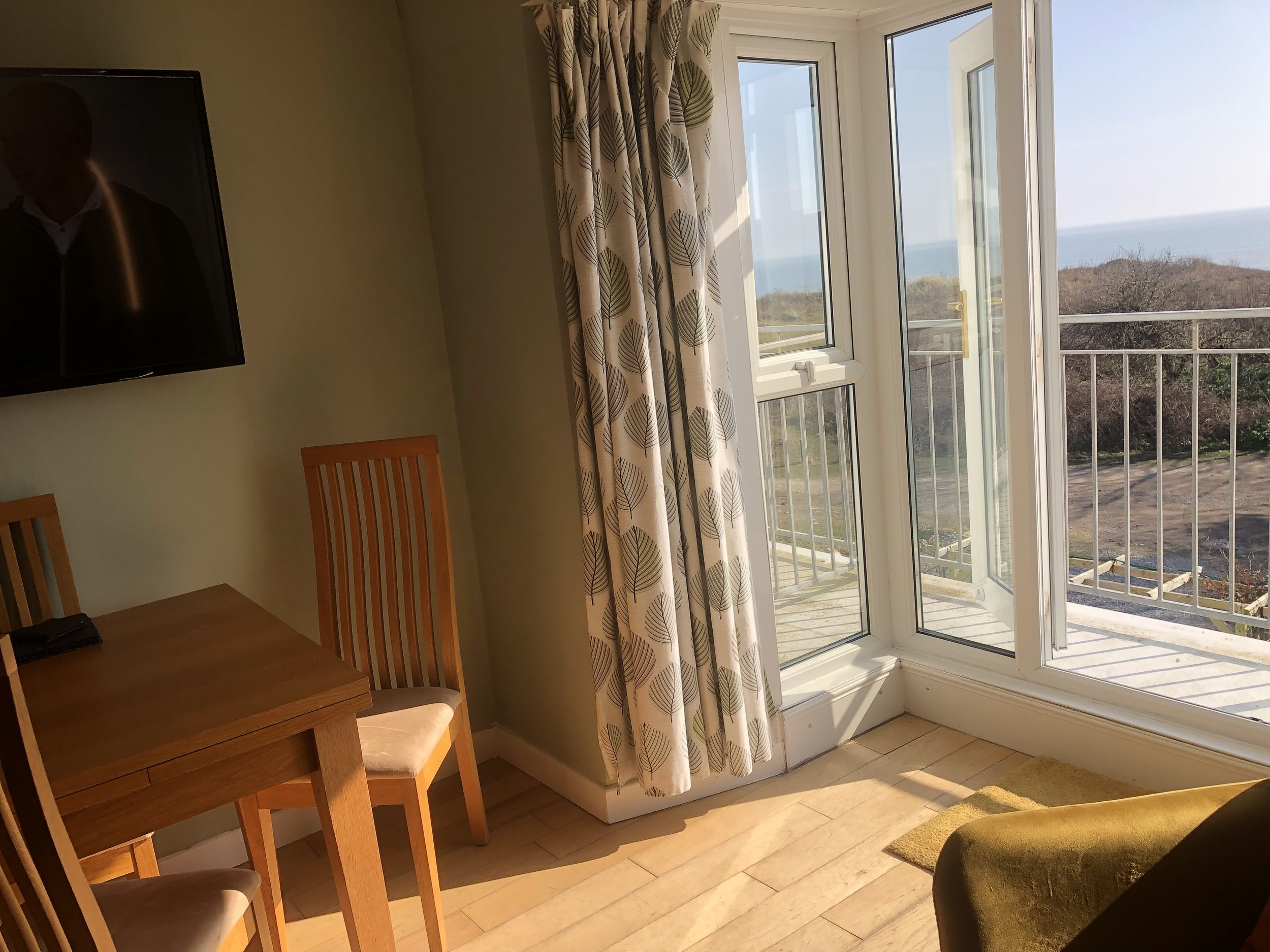 Room with wooden table and chairs, wall-mounted TV, and glass door opening to a balcony with a sea view.